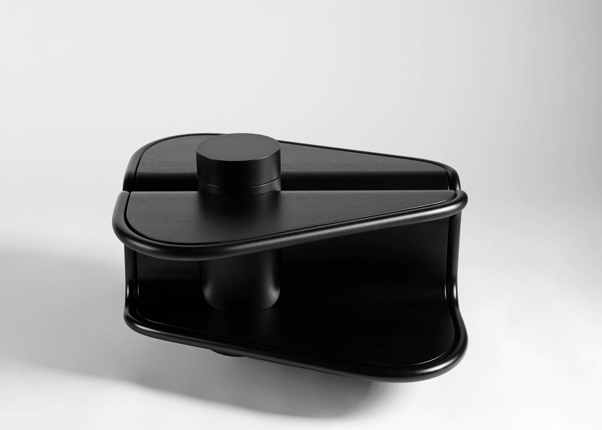 This image shows a side elevation view of Second Empress Coffee Table designed by Carol Egan.  The Ebonized mahogany table is shown against a grey background.  Upper and lower planes at opposing angles mirrored and intersected by a cylindrical form with swivel lid to reveal a concealed storage space.  Image shows the cylinder in the closed position.  The peripheral form of the coffee table is trimmed by a bisecting cylindrical edging.  The table is cantilevered from the upright cylindrical form to hover above the ground.  Dimensions of table are 26"w x 25"d x 16"h.