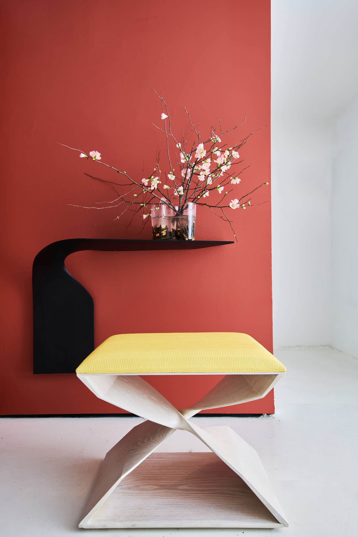 This image shows a x bench and a wall console both designed by Carol Egan and exhibited in her Decade show against a terracotta background.  The CE03 90° Twist Shelf in ebonized mahogany, 24"h x 36"w x 11.5"d (61 x 91 x 29cm). Wall-hung, cantilevered, shown here with an Alvar Alto vase and cherry blossoms.  In the foreground is an X bench in a cerused oak finish and custom quilted yellow leather upholstery.