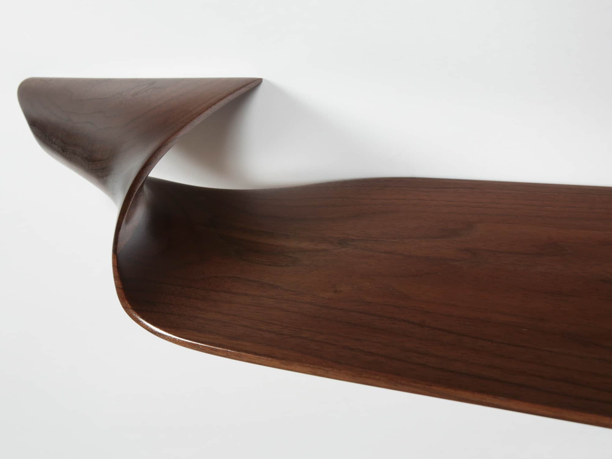 This image shows a detail of the end carving of the site specific CE16 Triple Twist Shelf in a walnut finish designed by Carol Egan.  The dimensions of the piece are 5.5"h x 64/58"w x 12"d (14 x 162/148 x 30cm). View Detail of end carving, wall-mounted and cantilevered around a corner.