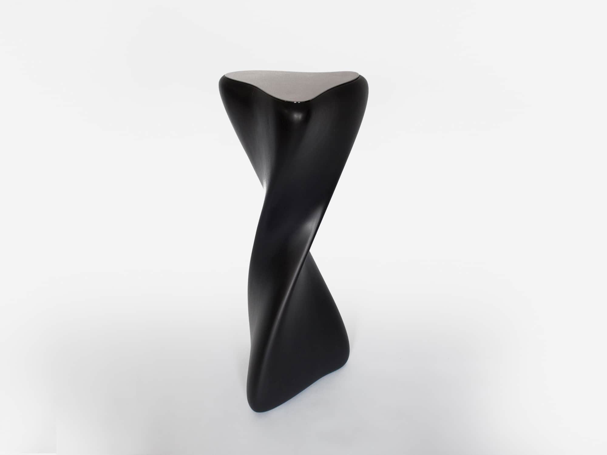 Designed by Carol Egan, this sculptural pedestal is shown here against a grey background.  The CE13 pedestal, 42"h x 16" diameter (107 x 40cm diameter) is in an ebonized mahogany finish and is a carved wooden pedestal with blackened nickel metal cap.