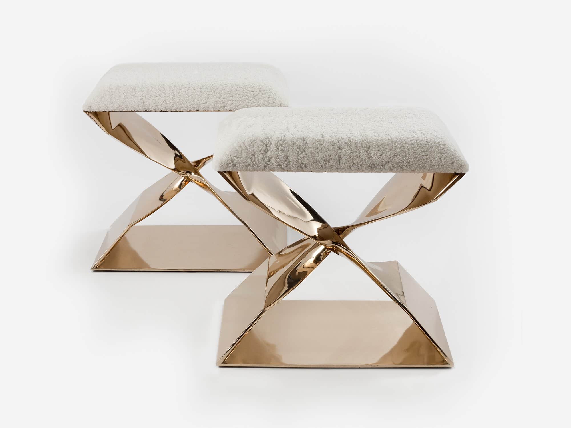 This image displays a pair of CE06 X- Stools in a polished Silicon Bronze finish designed by Carol Egan.   The dimensions of the stools are 18"h x 22"w x 15"d (46 x 56 x 38cm). They are shown in this image against a grey background with natural shearling upholstery.