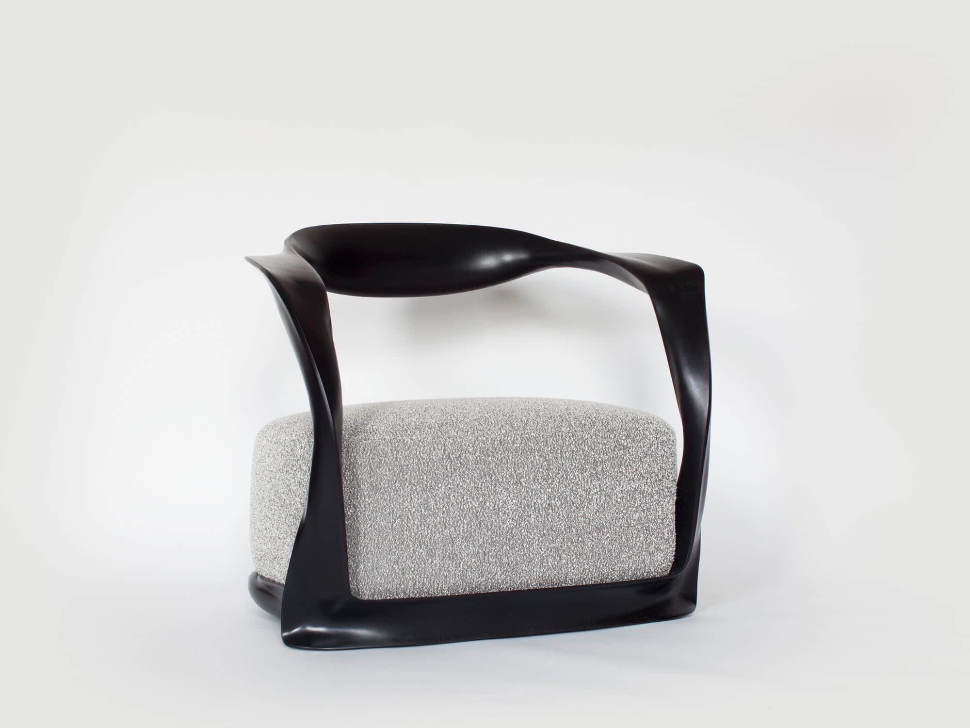 Fabricated in carved ebonized mahogany finish and shown here against a grey background is the CE15 Ombre armchair upholstered in a custom Silk boucle fabric designed by Sam Kasten.  The dimensions of the armchair are 28"h X 32"w X 24.5"d (70 X 80 X 60cm).  Designed by Carol Egan, the chair has a carved ribbon like frame with an open back and a sprung upholstered seat.