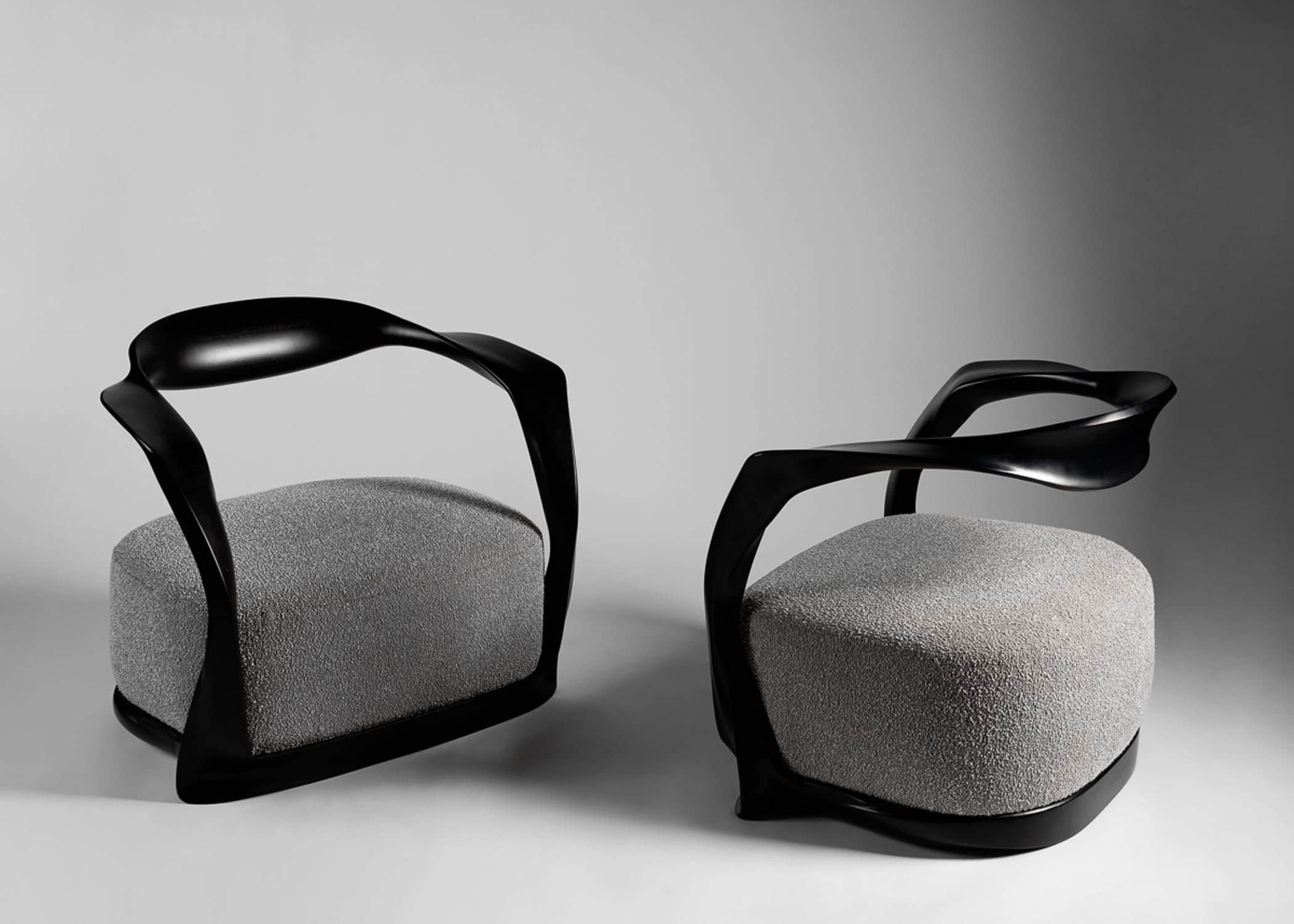 Fabricated in carved ebonized mahogany finish and shown here as a pair against a grey background are the CE15 Ombre armchairs upholstered in a custom Silk boucle fabric designed by Sam Kasten.  The dimensions of the armchair are 28"h X 32"w X 24.5"d (70 X 80 X 60cm).  Designed by Carol Egan, the chairs are carved in a ribbon like frame with an open back and a sprung upholstered seat.