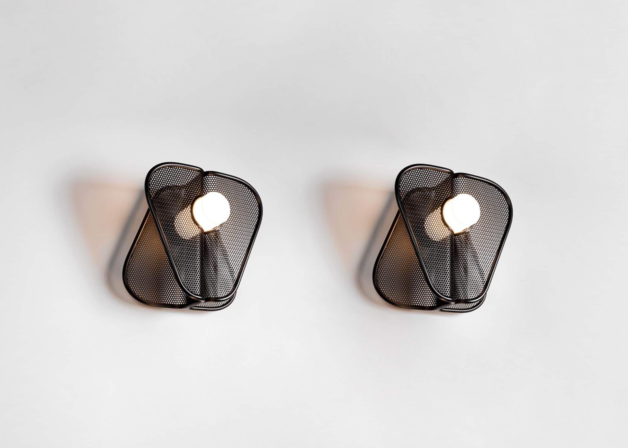 This image shows a pair of CE24 "Soverign" wall lights in blackened mesh and tubular metal frames designed by Carol Egan.  The wall light consists of opposing angled planes that are mirrored and intersected with a white tubular cylindrical acrylic led light. Shown here against a pale grey background the dimensions of the wall light are 14"h X 14"w X 9"d.