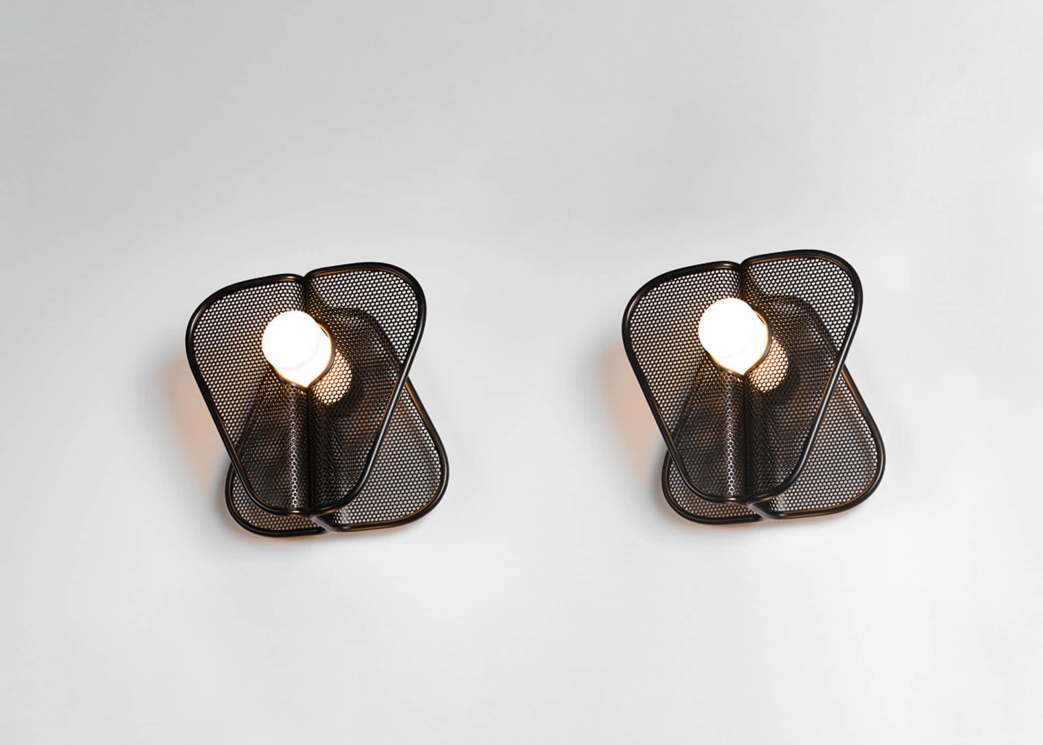 Designed by Carol Egan, a pair of CE24 "Soverign" wall lights in blackened mesh and tubular metal frames are shown against a grey background.  The wall light consists of opposing angled planes that are mirrored and intersected with a white tubular cylindrical acrylic led light. Shown herein a black and white finish, the wall lights are available in colored metal finishes and the dimensions of the wall light are 14"h X 14"w X 9"d.