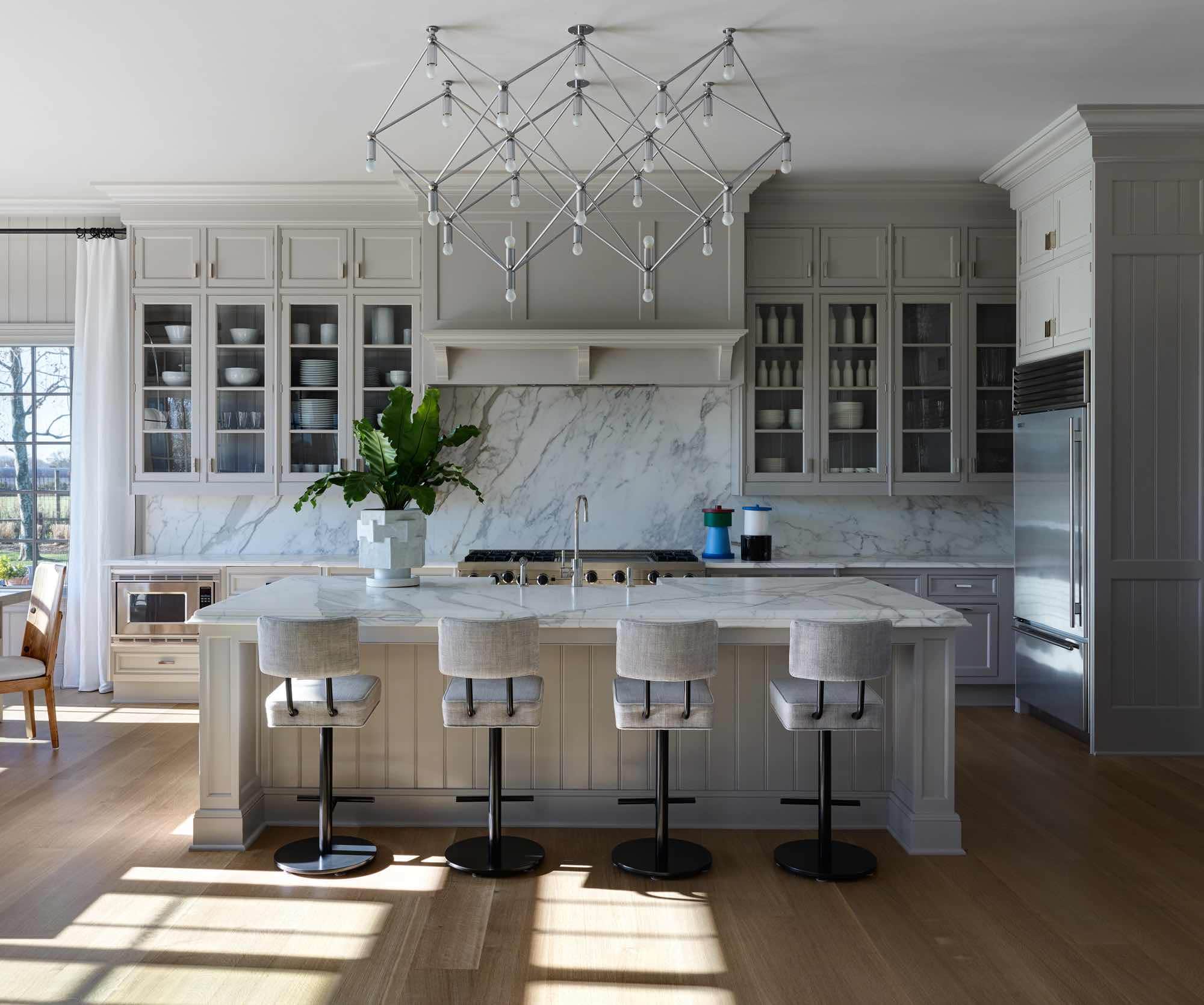 A photograph of a painted wooden kitchen with a statuary marble countertop flanked by custom barstools by Wyeth in Oscar Stone linen by C&C Milano.  Accents include Castagno & Tiglio ceramic vases with Lid by Nathalie du Pasquier and XL Marshmallow Hex planter by BZippy & Co.  Above the island, a sculptural light structure by Kinkeldey Studio from the 1970s lights the room.