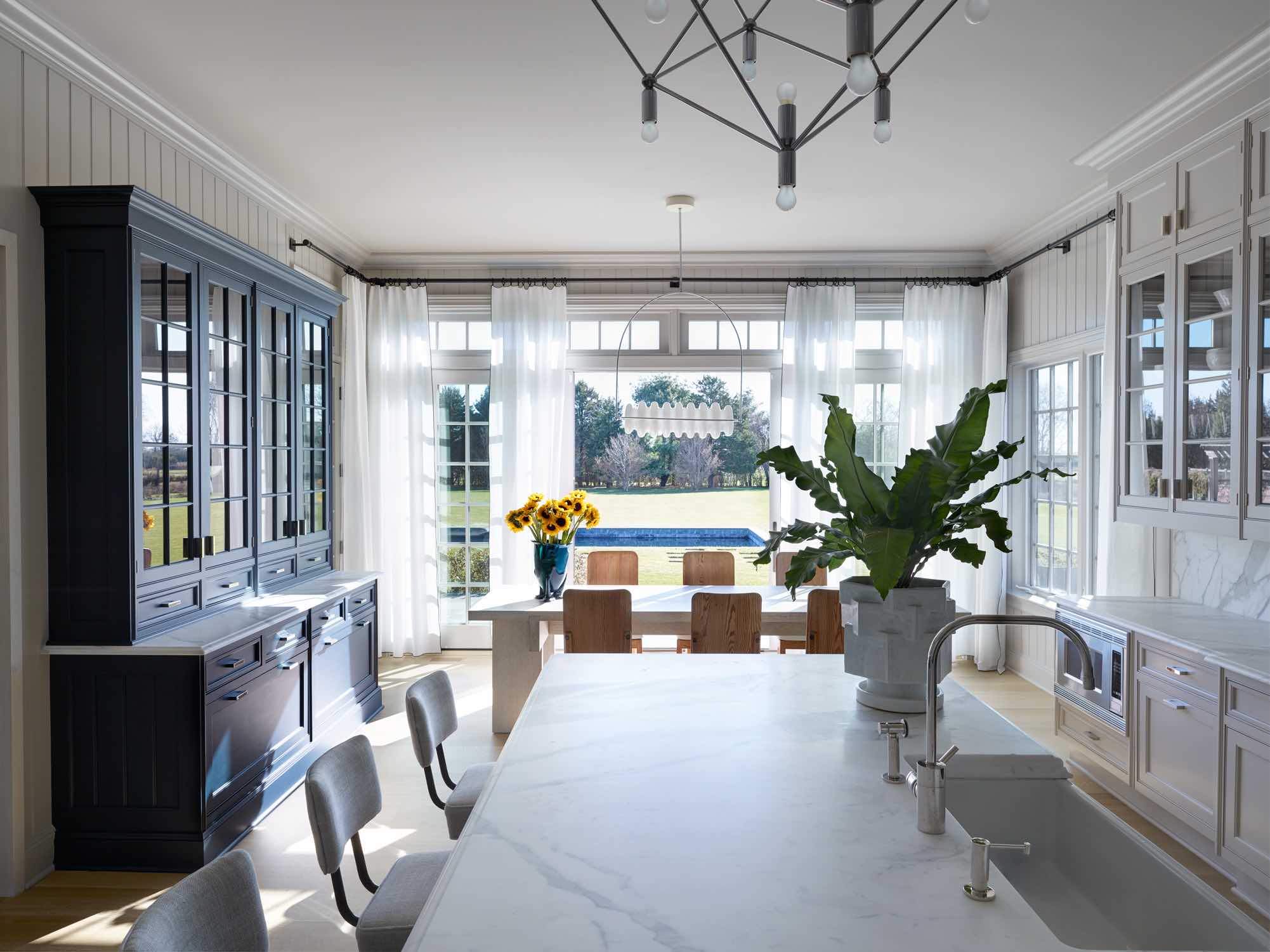 Shown here is an image of the kitchen and breakfast room area.  Jumbo Diamond vase by BZippy & Co. sits on top of the marble counter with Swivel bar stools by Wyeth.  Above the island is a 1970 light structure by Kinkeldey Studio.  A painted hutch cabinet contrasts the kitchen cabinet grey painted finish in an indigo finish.