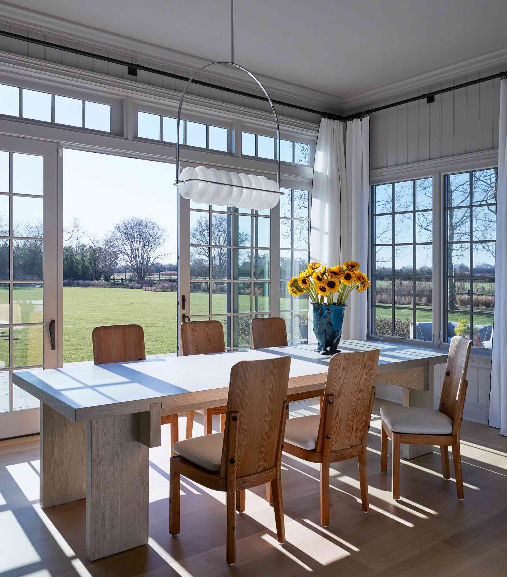 An image of the dining area in the kitchen designed by Carol Egan.  Lovö dining chairs by Axel Einar Hjorth surrounds the RAAF dining table by Piet Boon.  Sunflowers in Amazonia Resin vase by Gaetano Pesce accents the dining table.  Brucco hanging lamp by Éttore Sottsass lights the room.  Persico Mache linen curtains by C & C Milano lines the windows.