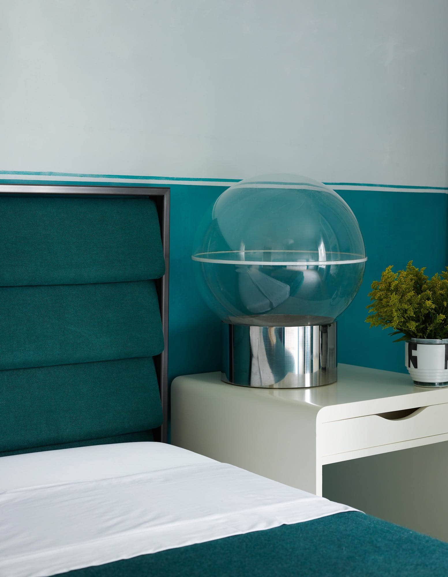 In this image is a guest bedroom designed by Carol Egan which consist of a custom headboard by Studio Carol Egan upholstered in Volterra Pino fabric by C&C Milano.  Beside is a "Trolley" Table by Jan Bocan with a vessel by Bodil Manz.  The wallcovering is two tone, teal and pale grey green hand-painted on canvas by Ann McGuire Studio.