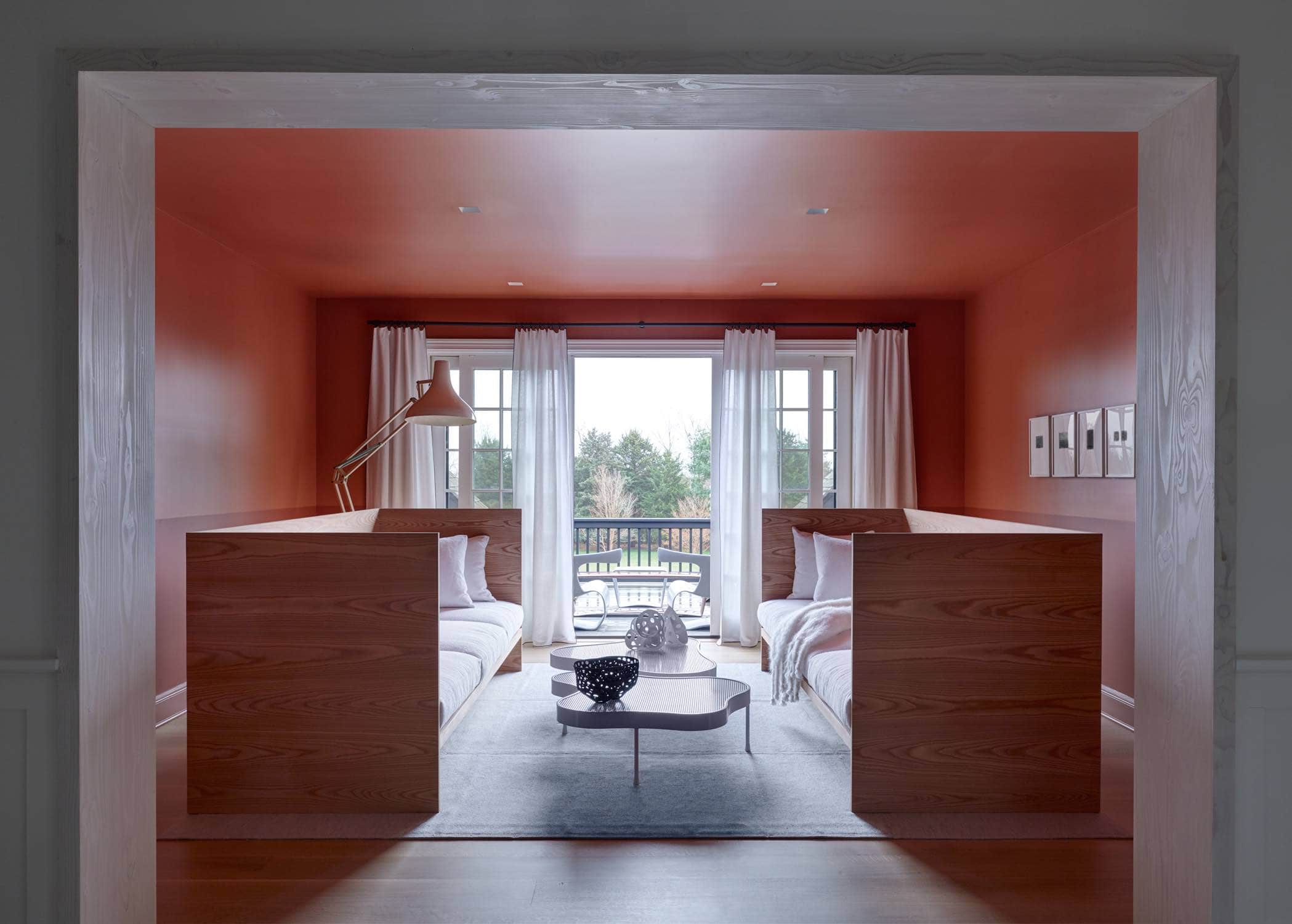 Designed by Carol Egan, this image shows a painted terracotta and plaster two tone sitting area with a pair of custom daybeds in Larchwood.  Between two daybeds are white perforated metal Biomorphic low coffee tables by Wyeth.  The walls are painted two-toned in Terre D Egypte and Modern Emulsion by Farrow & Ball.  A series of Photographs by Harry Callahan are framed in white painted frames and are hung horizontally above the line of the sofas.