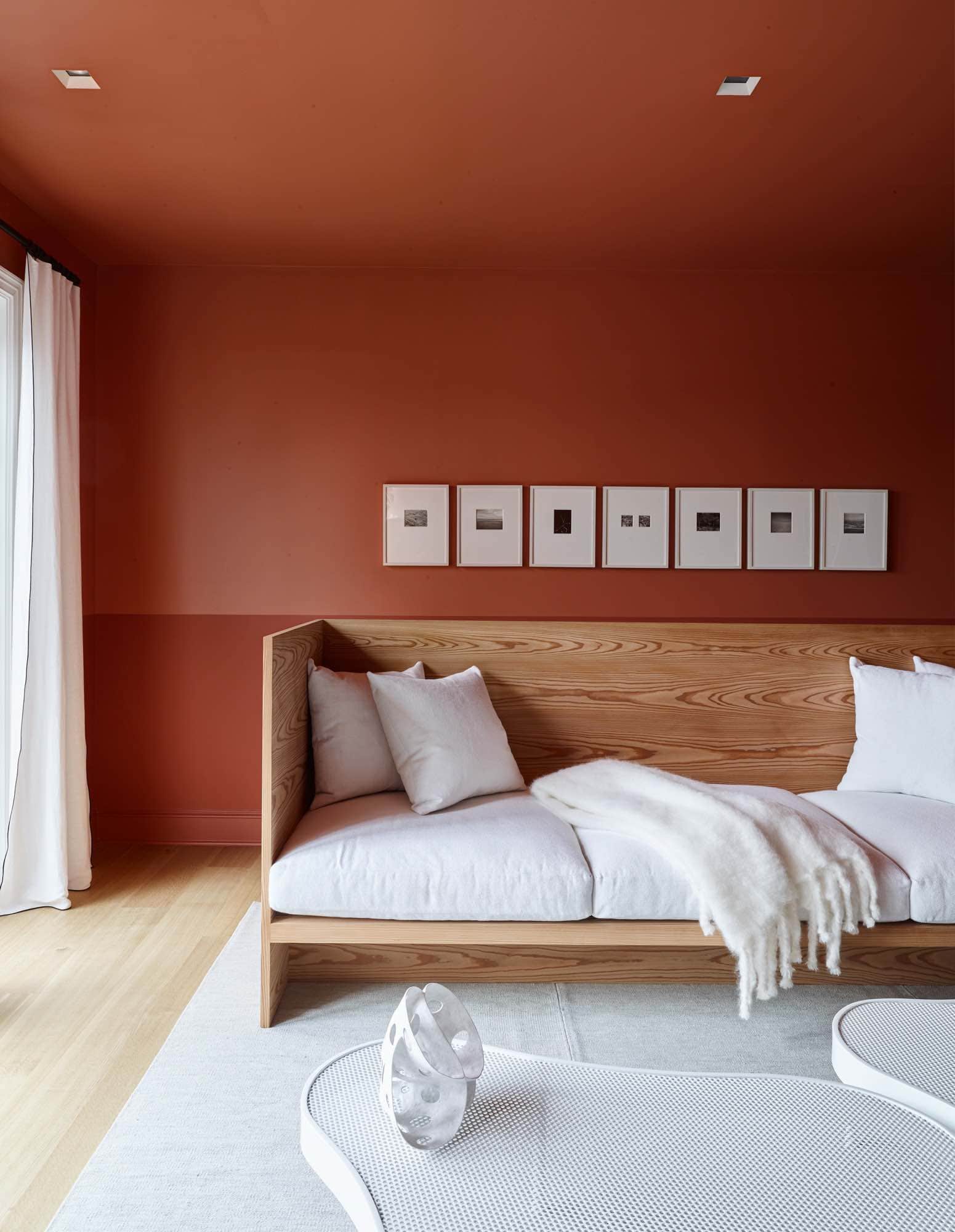 A photograph of a sitting area on the second floor of a Sagaponack residence.  It consists of a custom daybed in Larchwood with white cotton cushions and throw pillows shown with painted white perforated metal coffee tables by Wyeth. The windows are lined with Laveno Mache linen curtains by C&C Milano.  Two-toned walls in Terre D Egypte and Modern Emulsion by Farrow & Ball.  Photographs by Harry Callahan are hung horizontal in a series of painted white frames.  There is a Sam Kasten grey area rug in the room.