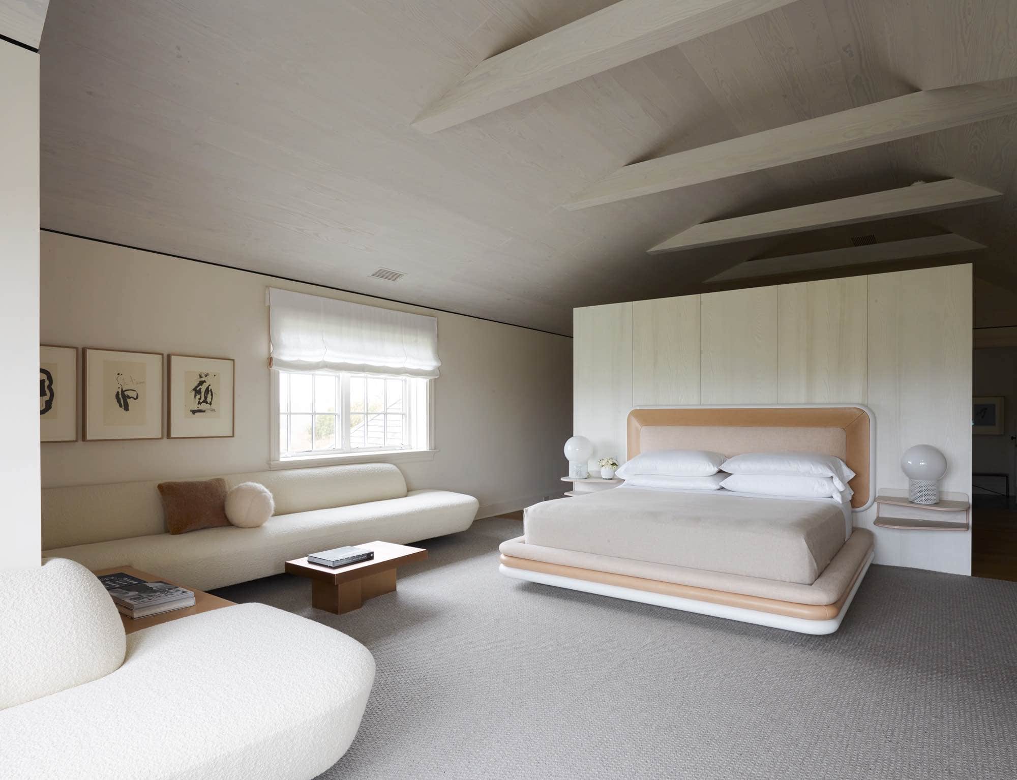 Shown here is an image of a master bedroom designed by Carol Egan.  The statement custom bed design by Studio Carol Egan is upholstered in old Rose Heathered Strie fabric by MM Design & ISEO IS16 leather by Foglizzo Leather.  Edition Verre Luminiere globe lamps by Michel Boyer sits on top of a pair of 1965 Bauhaus style wall hung shelves which flank each side of the bed.  The custom floating banquette design by Studio Carol Egan in Ivory Karakaroum fabric by Dedar Milano with ball cushion by Pierre Yovanovitch surrounding a coffee table 80 by Marcin Rusak is cantilevered around the corner of the room creating a long seating area.  Above the banquette is Octavio Paz Portfolio Lithographs by Robert Motherwell.