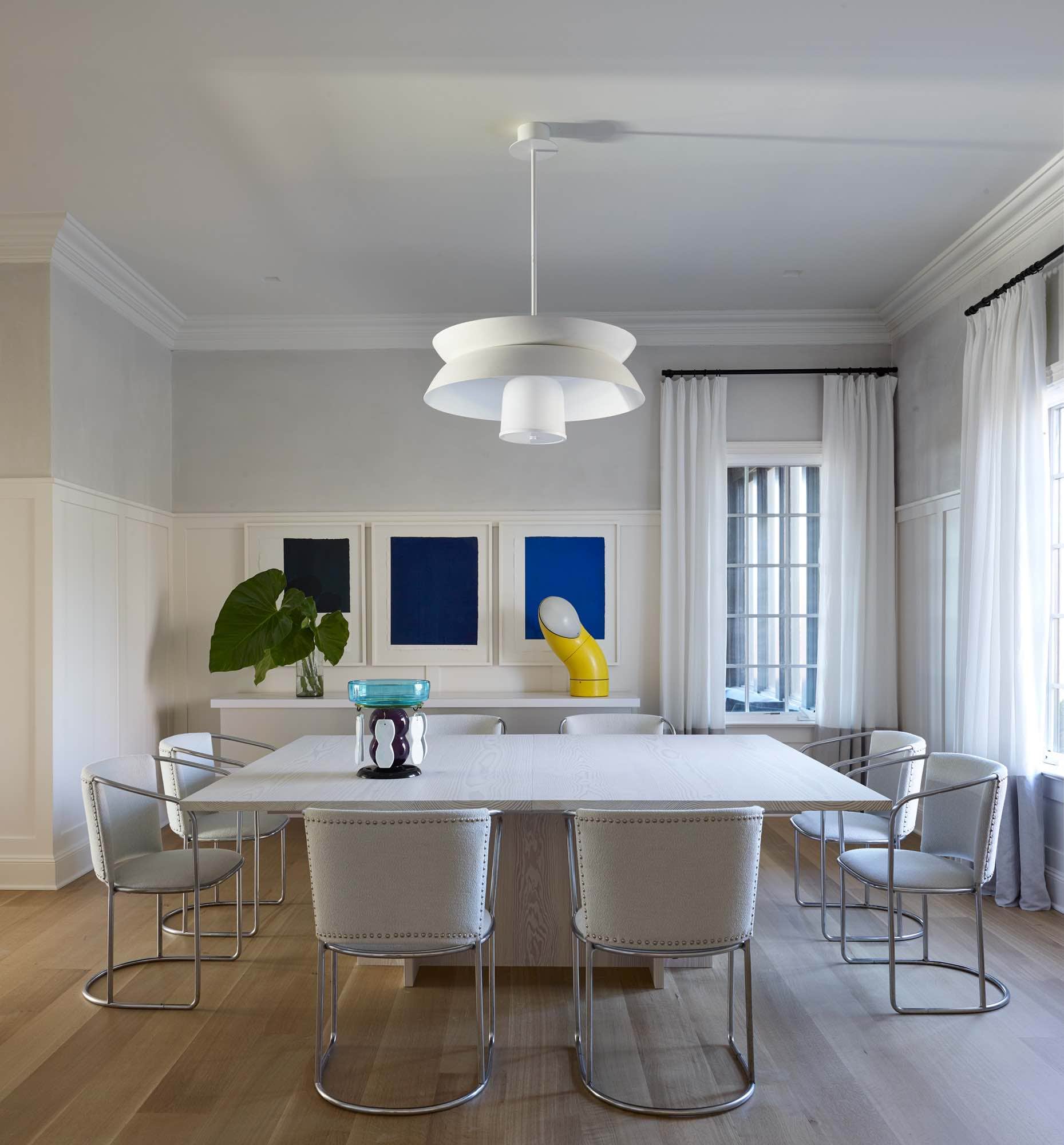 In this image is a dining room designed by Carol Egan.  The 1930s Tosse Bakery armchairs by Axel Einar Hjorth in mélange wool boucle in Brunswick Mirage by Holland & Sherry surrounds a custom wooden Doug fir dining table accessorized with a Filillia fruit basket by Ettore Sottsass.  The room is lit by a suspension Marceau pendant light by Eric Schmitt and a Tumbo lamp by Piero Menichetti.  “Untitled” Pastel on Two Rivers artwork by Callum Innes hang on the paneled wall above a wall hung console.