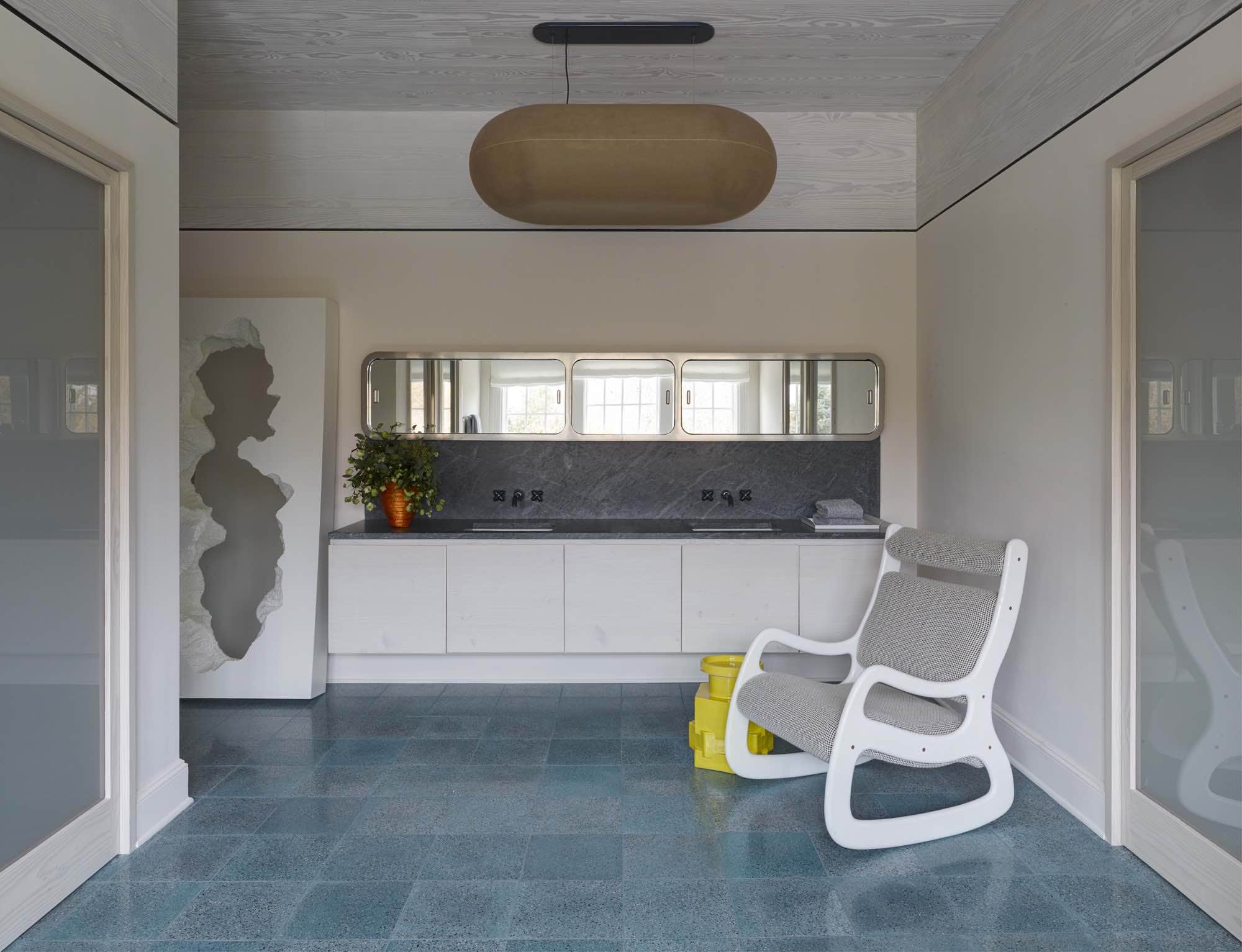 In this image of a master bathroom designed by Carol Egan, a rocking chair Gamme 1150 prototype by Jean Pierre Laporte beside a PLAXL in gloss yellow by BZippy & Co. sits on top of an Astra V103 Terrazo Field tiles in Agate blue by Urban Archaeology floor.  The statement Puffball pendant by Faye Toogood hangs on the ceiling.  The wall hung vanity has a soapstone counter and backsplash and an oval framed metal and mirror pill shaped medicine cabinets hanging above.  The ceiling is planked with Dinesen Douglas Fir and the walls have a painted plaster finish.