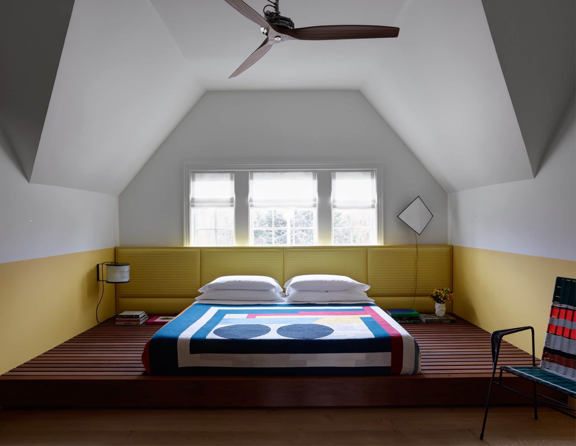 Shown here is an image of a guest bedroom designed by Carol Egan with a low Ipe wooden platform that houses the mattress on a low platform.  The yellow leather and painted wood headboard are wall hung above the platform bed and upholstered in a custom Zig Zag quilted leather by Capricorn Hides.  The bed is dressed in a commune quilt by Adam Pogue in bright geometric patterns.  A Serie Fresnel 1139 sconce by Joe Colombo and a 1970s Angular chandelier hang on the bedside walls.  Armchair by Marni Market.