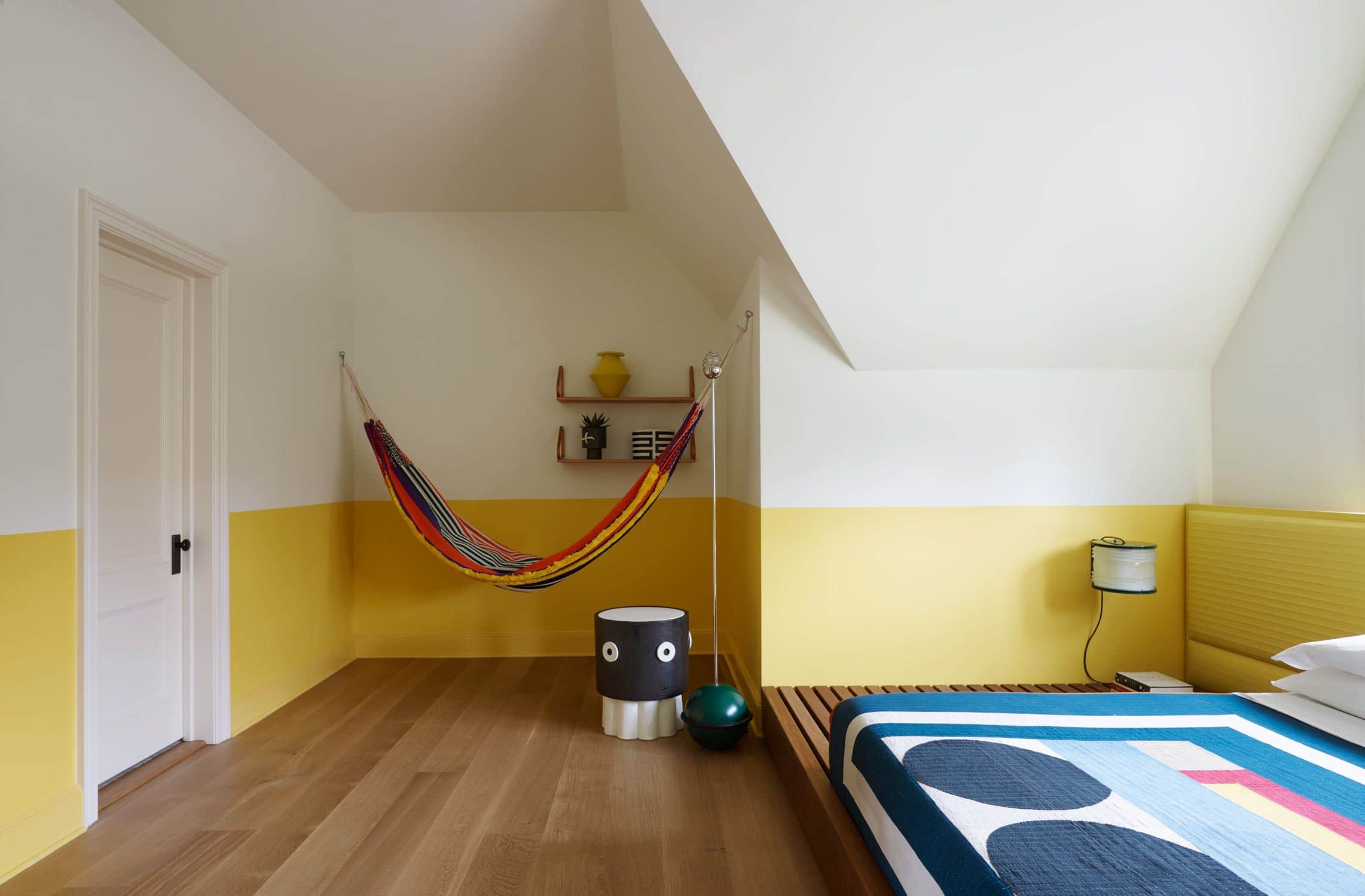 Designed by Carol Egan, this image shows a guest bedroom painted in bright yellow and white with a custom designed platform bed fabricated in Ipe wood.  Hung wall to wall in the corner of the room is a multi-colored woven hammock from Marni. The bed is dressed in a multi-colored geometric patterned Commune quilt by Adam Pogue.   Above the hammock are wall shelves by Alva Aalto and a side table in black and white ceramic.