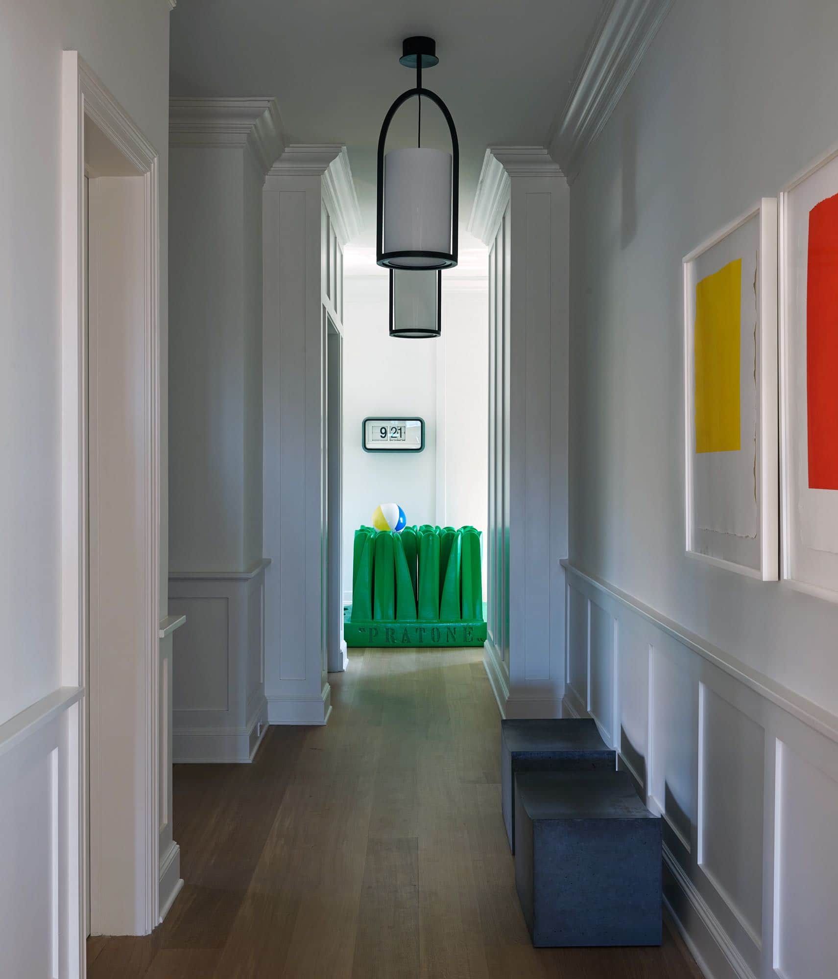 Shown here is an image of a hallway off of the entry foyer with paneled walls, painted in white and plank oak wood flooring.  At the end of the hallway is a Pratone chaise lounge by Ceretti, Derossi, & Rosso for Gufram.  The hallway designed by Carol Egan has two benches along the hallway under the color blocked Callum Innes prints which are hung above the Wainscott panel.  the benches are cement pillows by Chris Lehrecke.  There are a series of pendant ceiling lights in bronze and white opaline glass that are hung in the hallway designed by Eric Schmitt