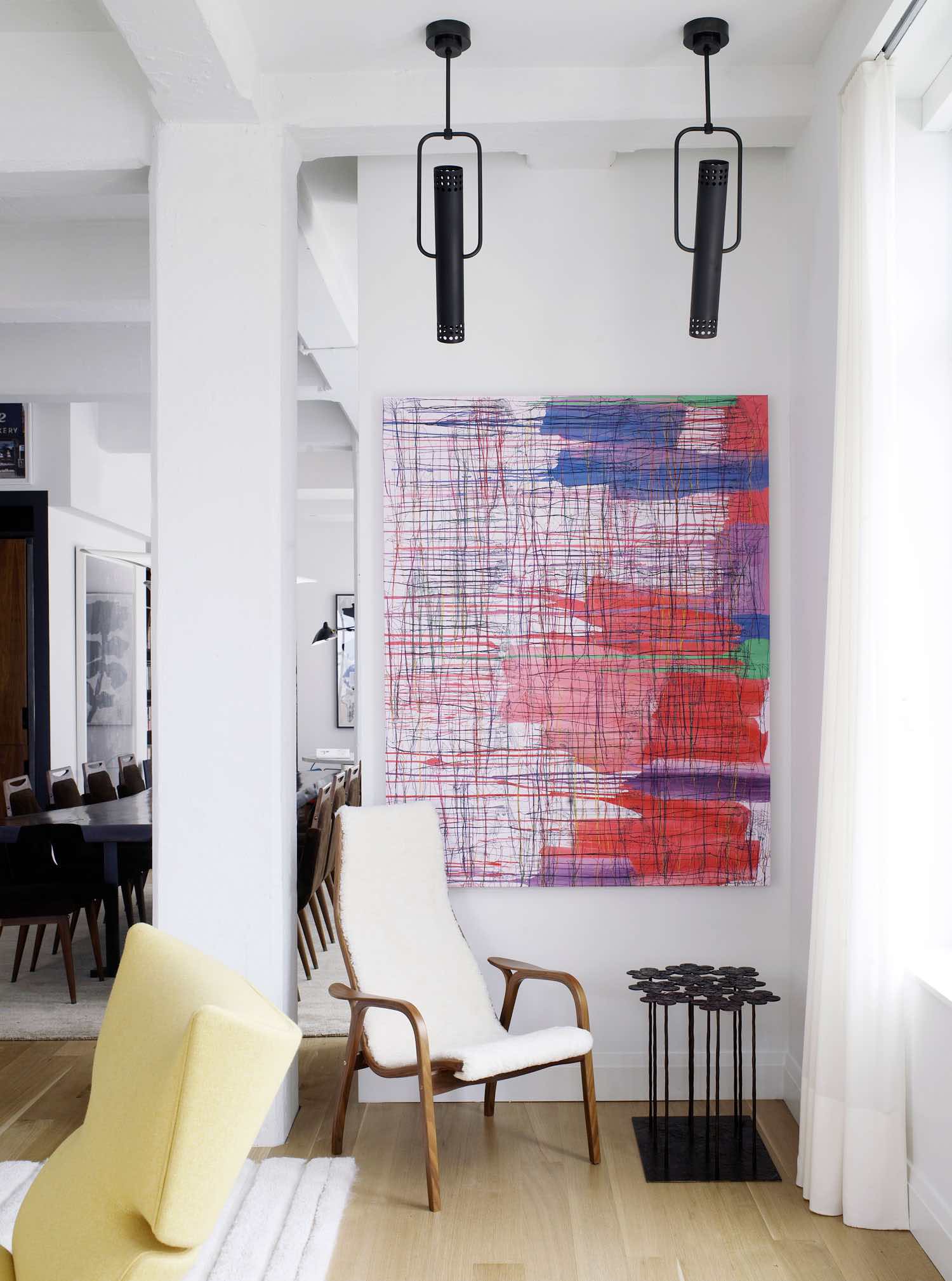 Featured in this image is an artwork by Ghada Amer in acrylic and embroidered thread hanging in a living room designed by Carol Egan in a Tribeca Loft.   A Lamino Chair by Yngve Ekström upholstered in natural shearling sits next to a side table in bronze by Hubert Le Gall under the artwork.  Flooring is wide plank oak, and the kitchen and dining area can be seen in the background.