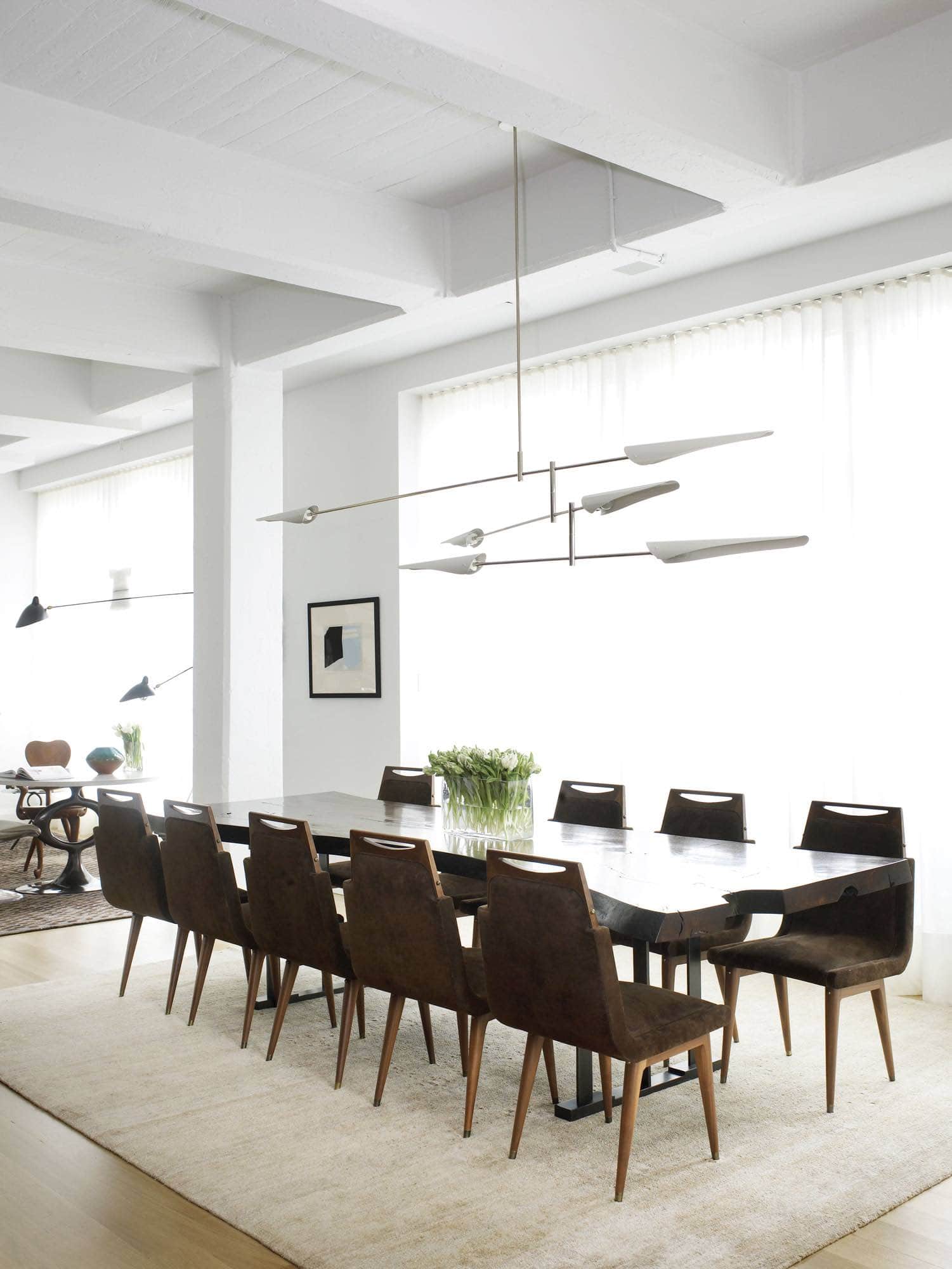 This image shows a view of the dining room designed by Carol Egan featuring a slab live edge walnut table with cast bronze base flanked with vintage Brazilian dining chairs with rosewood frame and upholstered in chocolate brown suede.  Above the table a Sarus three-tier chandelier by David Weeks hangs and the room is anchored by a nettle and silk area rug in camel tones.