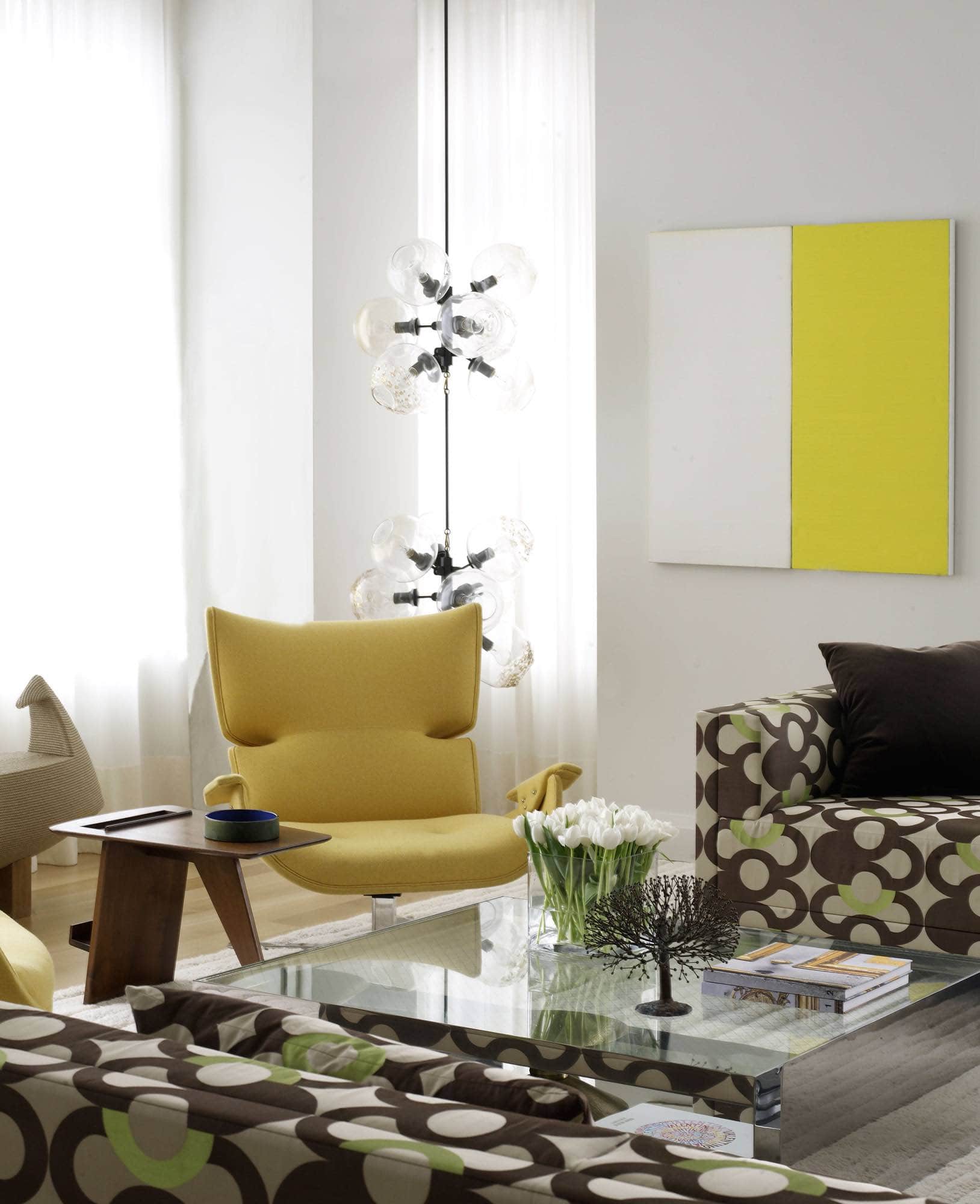 Designed by Carol Egan, this image shows the living room in a Tribeca loft.  The "At One Remove" painting on canvas by artist Callum Innes anchors the room.  A pair of custom sofas designed by Studio Carol Egan in Prelle fabric designed by House of Leleu in the 1950s are a graphic contrast in the room.  Flanked by a pair of Paulistana lounge armchair by Jorge Zalszupin and upholstered in a vibrant yellow wool.  A rope "Piri" Bench by Christian Astugevieille floats below the large loft windows.  The coffee tables, a pair by Joe D’Urso are industrial glass a steel.  A light sculpture by Lindsey Adelman hangs in the corner of the room.