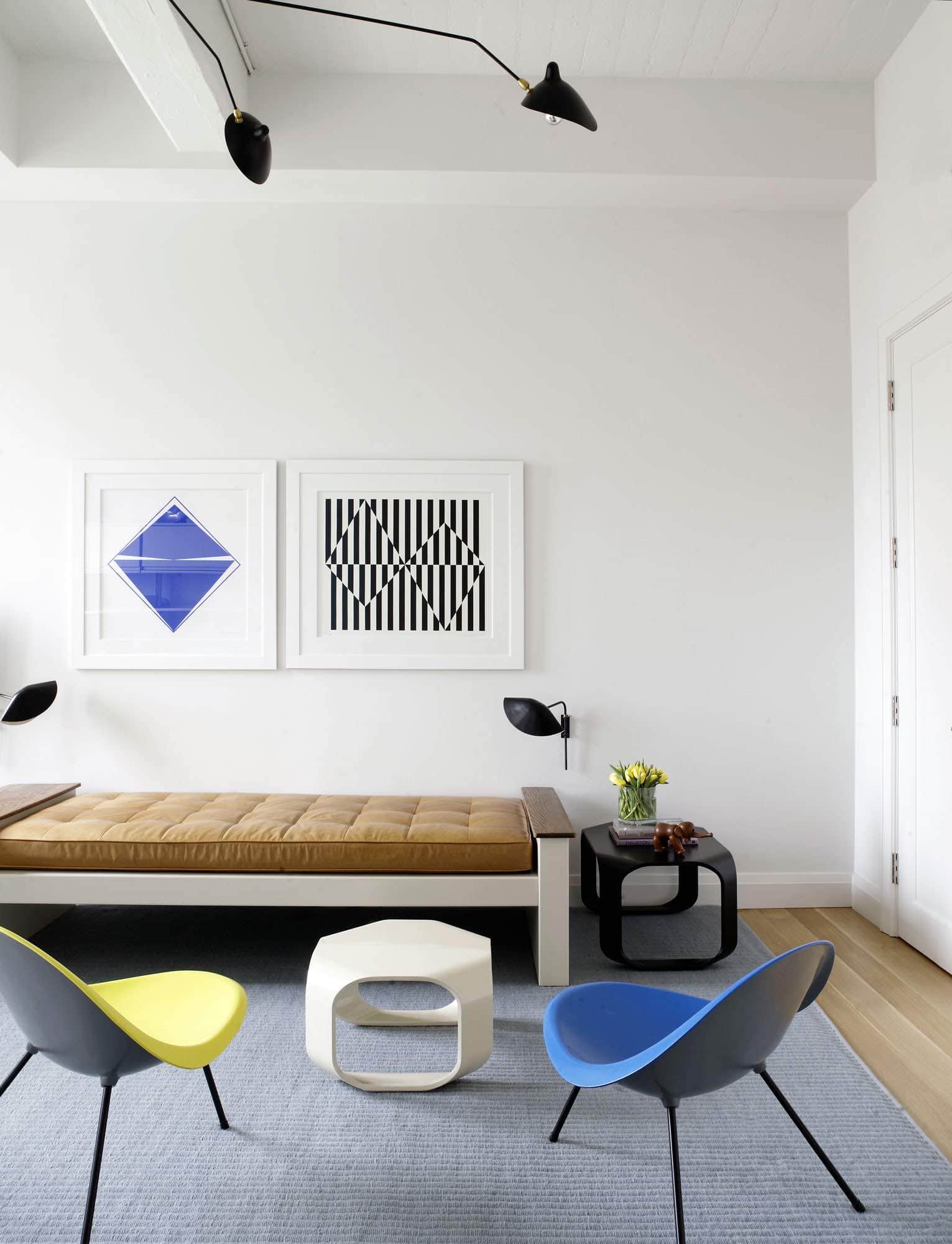 This photograph shows a view a study designed by Carol Egan in a Tribeca loft.  With two aluminum tripod chairs by Poul Kjaerholm opposite a Jean Prouve Metal Daybed. A serge Mouille Three arm light fixture hangs from the ceiling in blackened Metal and a pair of graphic modernist prints are framed on the wall above the day bed by Carmen Herrera.  A handwoven blue green looped area rug can be seen against the oak flooring in the room.