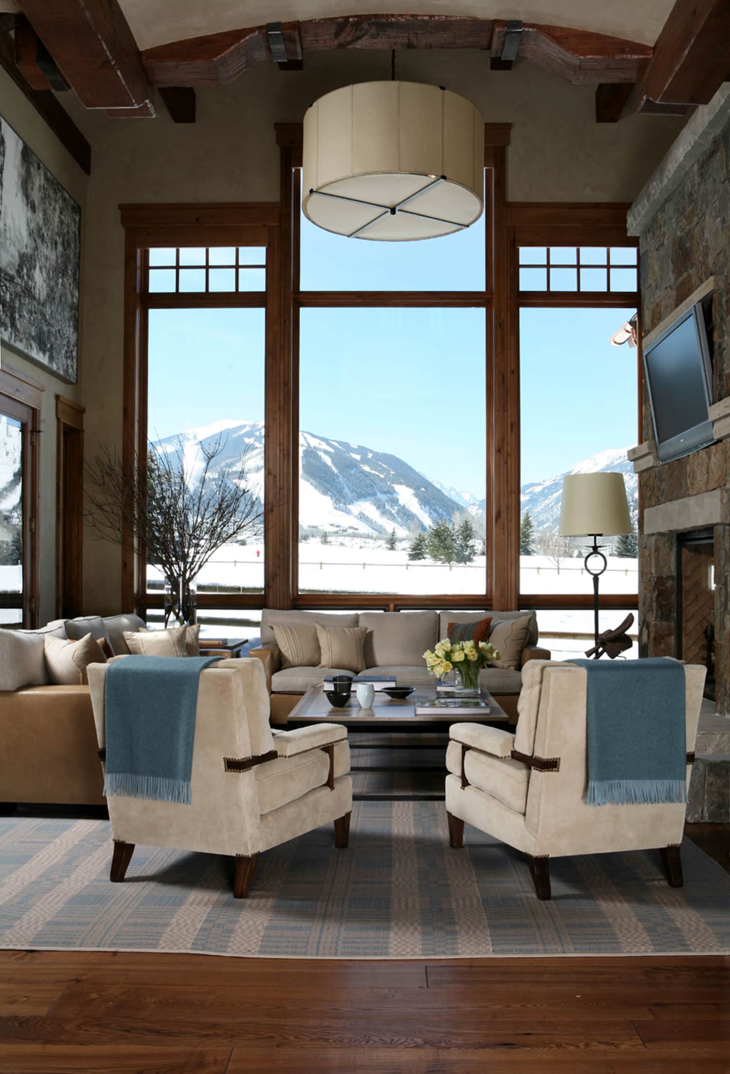 Designed by Carol Egan, this image shows a double height living room in an Aspen residence with large windows overlooking snowcapped mountains.  A large vellum shade Pendant light fixture hangs above the seating area in the living room.  Two Armchairs by Roman Thomas are upholstered in corduroy fabric and sit on a handwoven area rug in linen and pale blue wool.