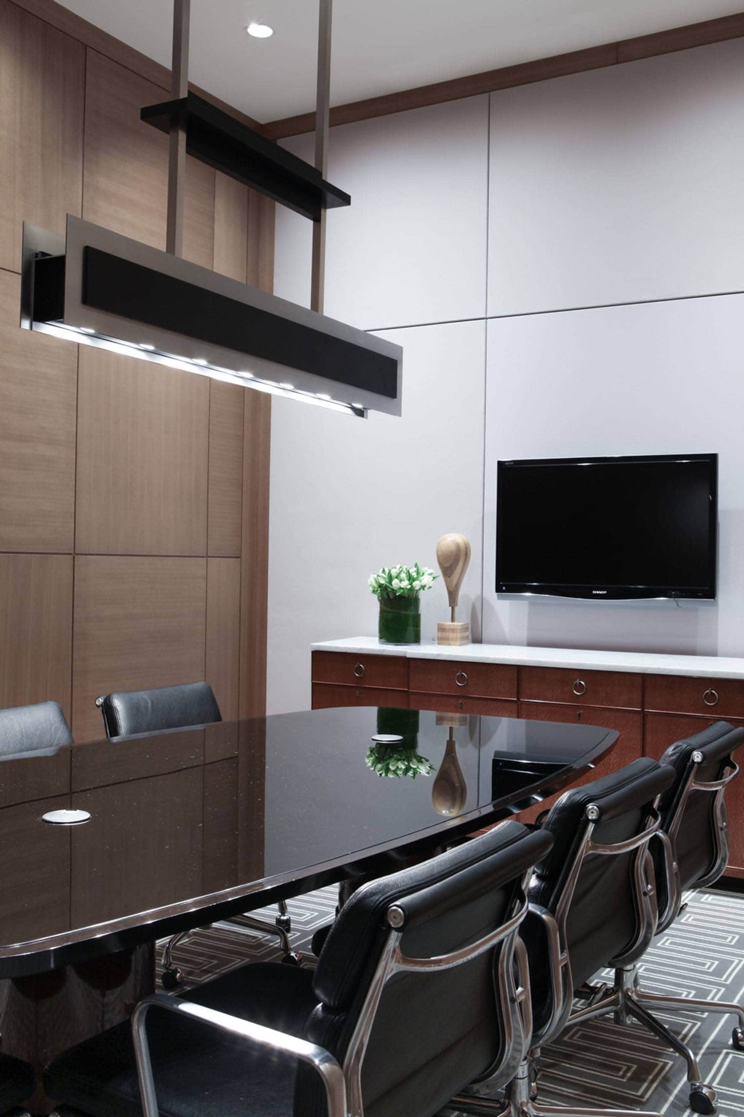 Designed by Carol Egan, this oak paneled executive conference room shows a custom black-painted and satin aluminum linear ceiling lamp hanging over the black opaline glass and walnut bow shaped conference table.  Soft pad management chair by Charles and Ray Eames flank both sides of the conference table.  Custom handwoven wool area rug in pale grey and Greek key border Motif.