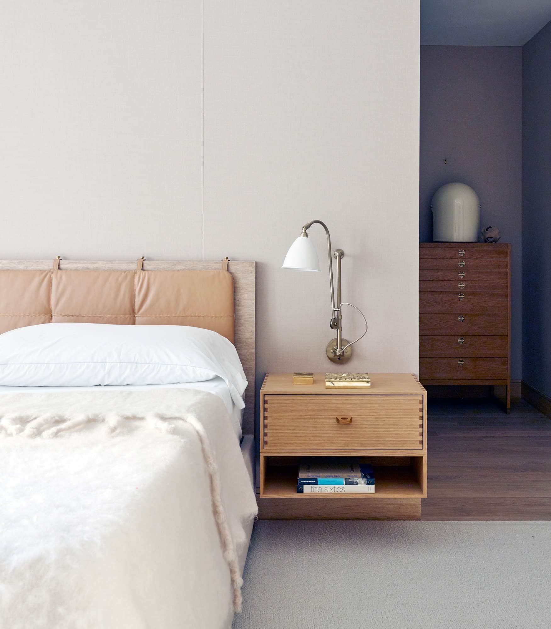 This image shows the master bedroom which has upholstered camel flannel wallcovering and a custom designed bed by Studio Carol Egan. Headboard cushion in classic Iseo by Foglizzo Leather & glazed canvas in biscuit by MM Design Fabrics. You can see wall hung night tables in Oak flanking the bed.  Installed over the bedside for reading are a pair of BL5 wall lamps by Robert Dudly Best Alpaca area rug by Mitchell Denberg in the Master Bedroom.