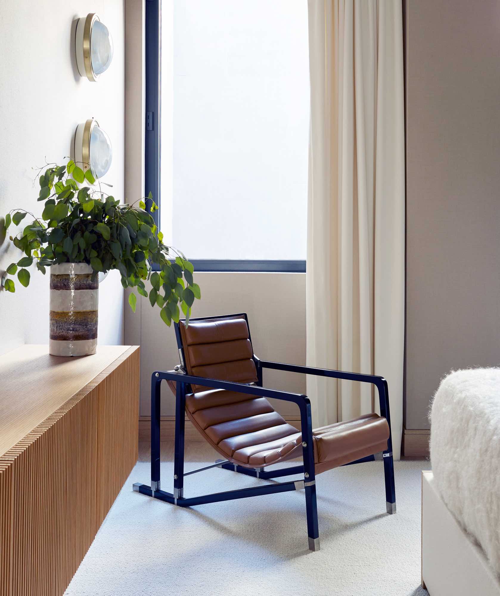 This image shows a detail shot in the master bedroom.  Nestled in the corner by the window is the iconic Transat chair by Eileen Gray.   A custom wall hung oak cabinet with ribbed detailing along the doors is visible in the foreground.  There is an Alpaca area rug by Mitchell Denberg in the room.  There are three wall lights by Sergio Mazza stacked one on top on another and wall mounted by the armchair for task lighting.  Curtains in bechamel stork by Rogers & Goffigon are tailored and asymmetrically stacked by the window.