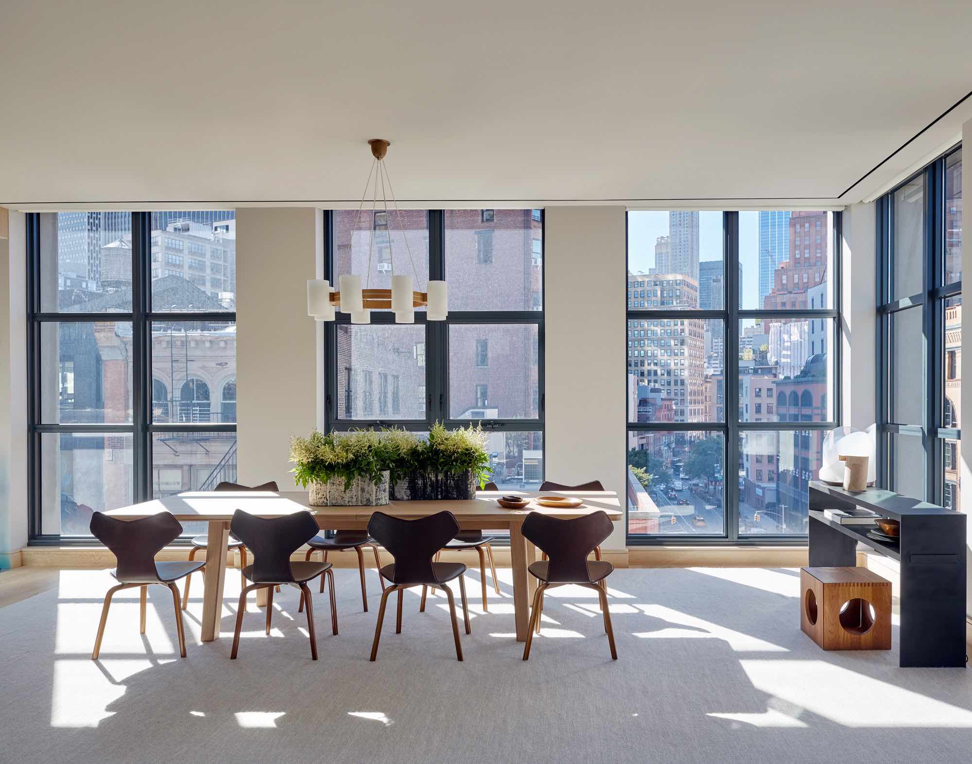 In the same loft apartment, this image reveals the loft dining room area with double exposure full length windows overlooking downtown Manhattan. A Luxo chandelier by Uno & Osten Kristiansson hangs over the oak dining table with Grand Prix chocolate brown leather dining chairs by Arne Jacobsen.  There is a handwoven area rug in heathered grey colors with a Custom blackened steel console by Eric Slayton which acts as a server to the dining table.  on the console is a Carlo Nasan Murano glass sculptural table lamp.  On the dining table is a beautiful ceramic planter by Kathy Erteman.