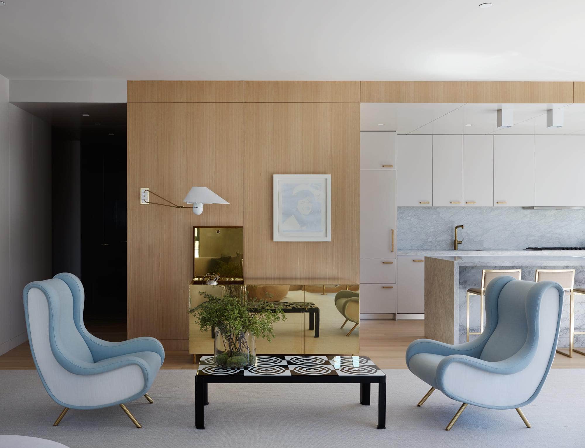 Shown in this image is the oak paneled living room area detailing a Brass bar cabinet designed by Ilse Crawford.  also visible is the panel ceiling of the kitchen which is inset in a painted Dior Grey Finish with brass hardware accents and a Cararra Marble Island.  There are two Vintage Señor chairs by Marco Zanuso flanking a Custom "Optical" coffee table by Roberto Rida. Hand-woven wool maresia area rug by Mitchell Denberg.