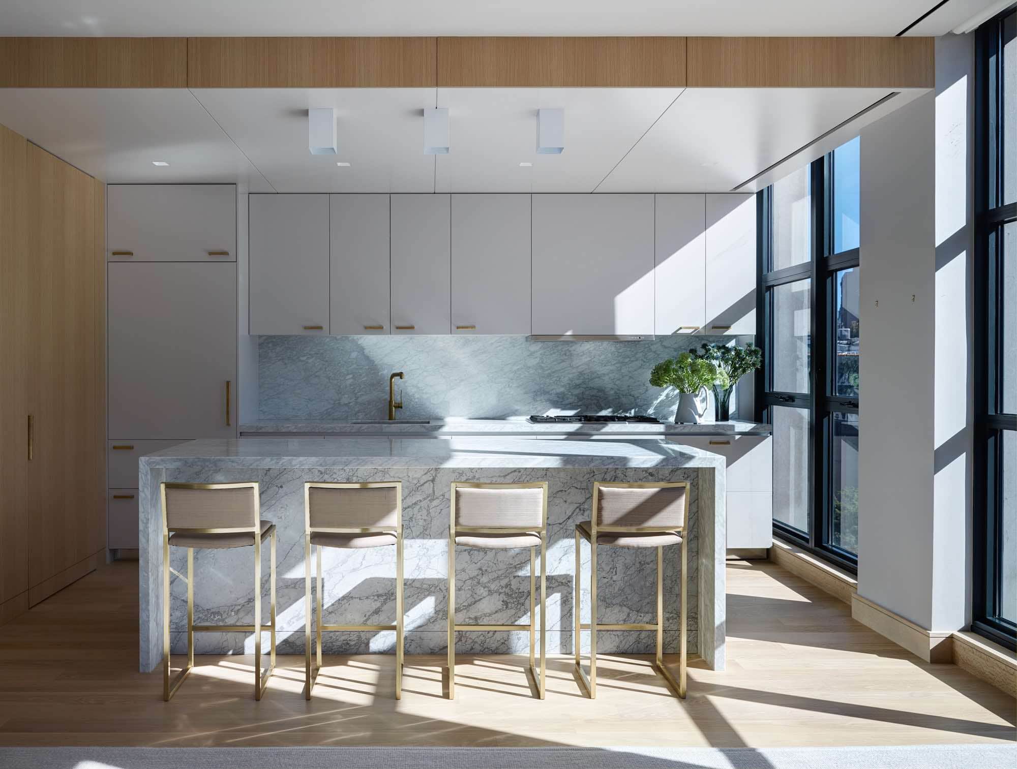 Vintage barstools by Willy Rizzos studio in Paris flank the Kitchen Island of Carrara Marble in this image of the loft designed by Carol Egan Interiors.  The paneled kitchen ceiling houses three Ceiling light fixtures by RAAK.  There is a composition of a blown glass vase by Per Lütken and a Ceramic pitcher by Astier de Villatte with flowers on the kitchen countertop.  The architectural hardware is by E.R. Butler & Co. and is in a radial bronze polished finish.