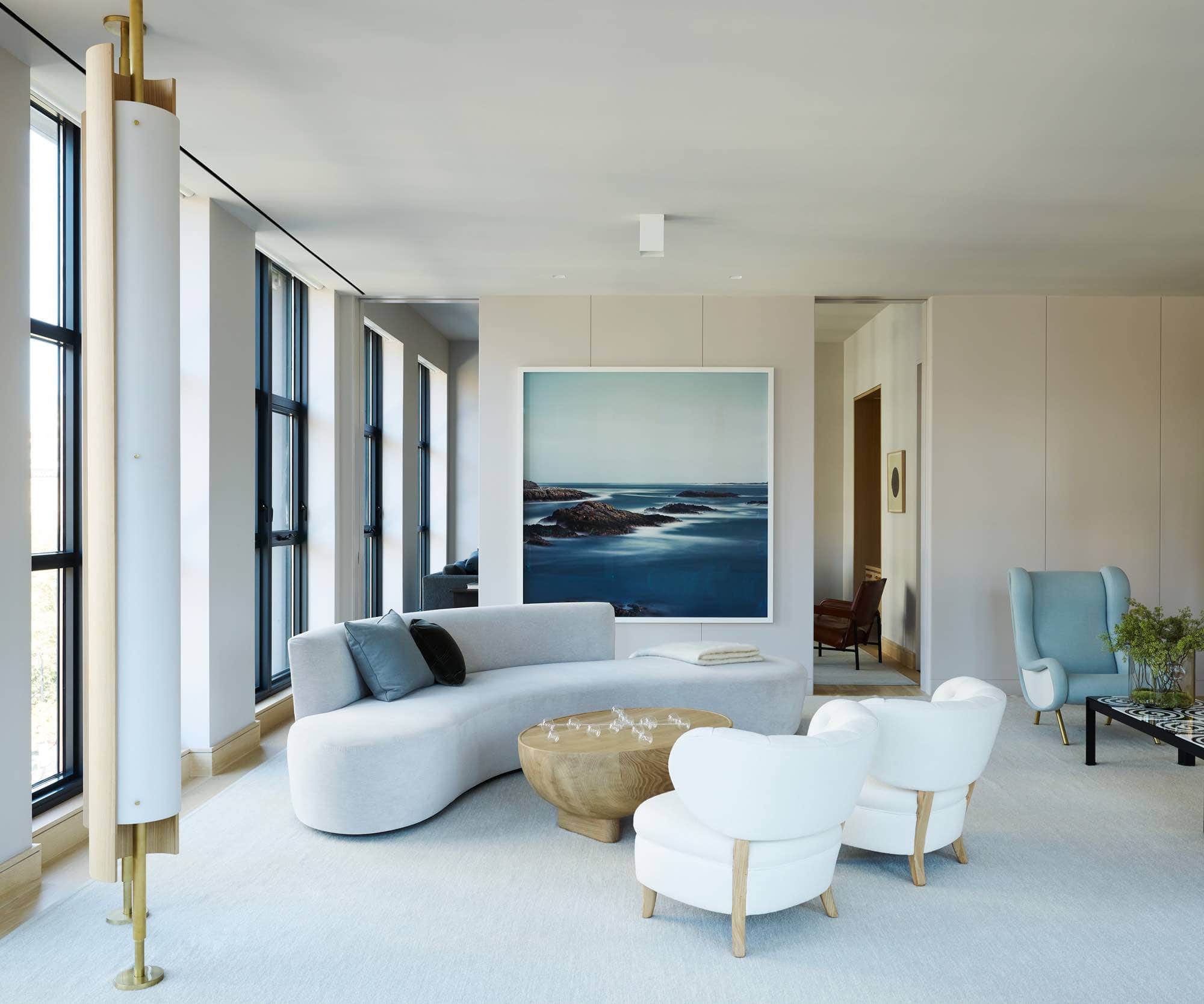 This image shows another angle of the living room area in the loft focusing on a large artwork that is titled ‘Fullmoon at Malin Head’ by Darren Almond.  The Artwork depicts a seascape and is hung on the living room wall behind a serpentine shaped custom sofa design by Studio Carol Egan sits opposite a pair of Vintage lounge chairs by Otto Schulz with a sculptural Oak coffee table.  There are two Oak and Brass Pillar lights floor to ceiling designed by Studio Carol Egan in the foreground.