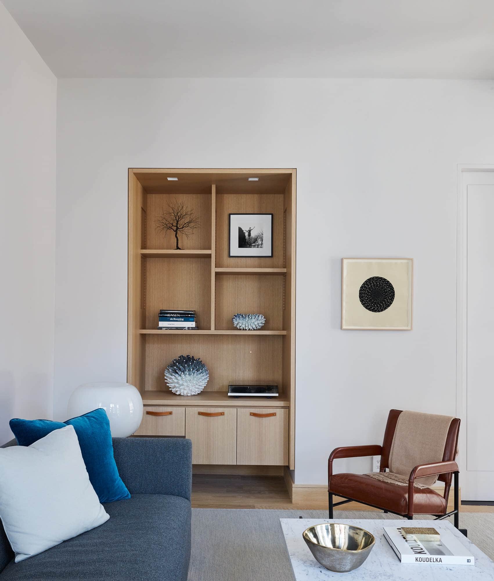 This image shows a custom designed bookcase inset into the wall by Studio Carol Egan located in the Family TV room.  Objects are style on the oak bookcase like a Wire tree sculpture by Pablo Avilla, Ceramic sculptures by Eva Zethraeus.   There is a JMF style Sofa in a grey boucle fabric and vintage leather French Armchair from the 1960s.  On the Steel Sofa end table, you can see an Arteluce translucent glass table lamp.  the coffee table in the room is in the manor of Poul Kjaerholm by Jonathan Burden Gallery with Rupi nesting bowl by Jaimal Odedra as accessories on the tabletop.