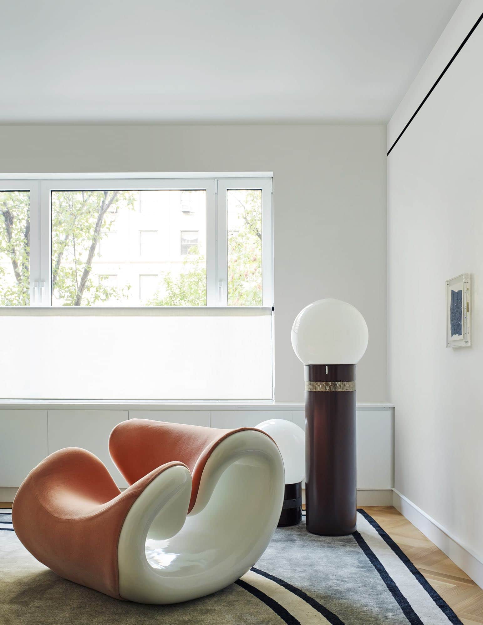 This photograph shows a detail shot from a Park Avenue living room designed by Carol Egan.  There is a 1970s Esox Chair by Jean-Pierre Laporte, experimental and sculpturally formed chair in white fiberglass frame with a powder pink Mohair upholstery.  Next to this playful chairs are Orocolo floor lamps by Gae Aulenti, one tall, one low.  An artwork by Gabriel de la Mora called Collage: "Ultramarine Blue" is hung on the adjacent wall.