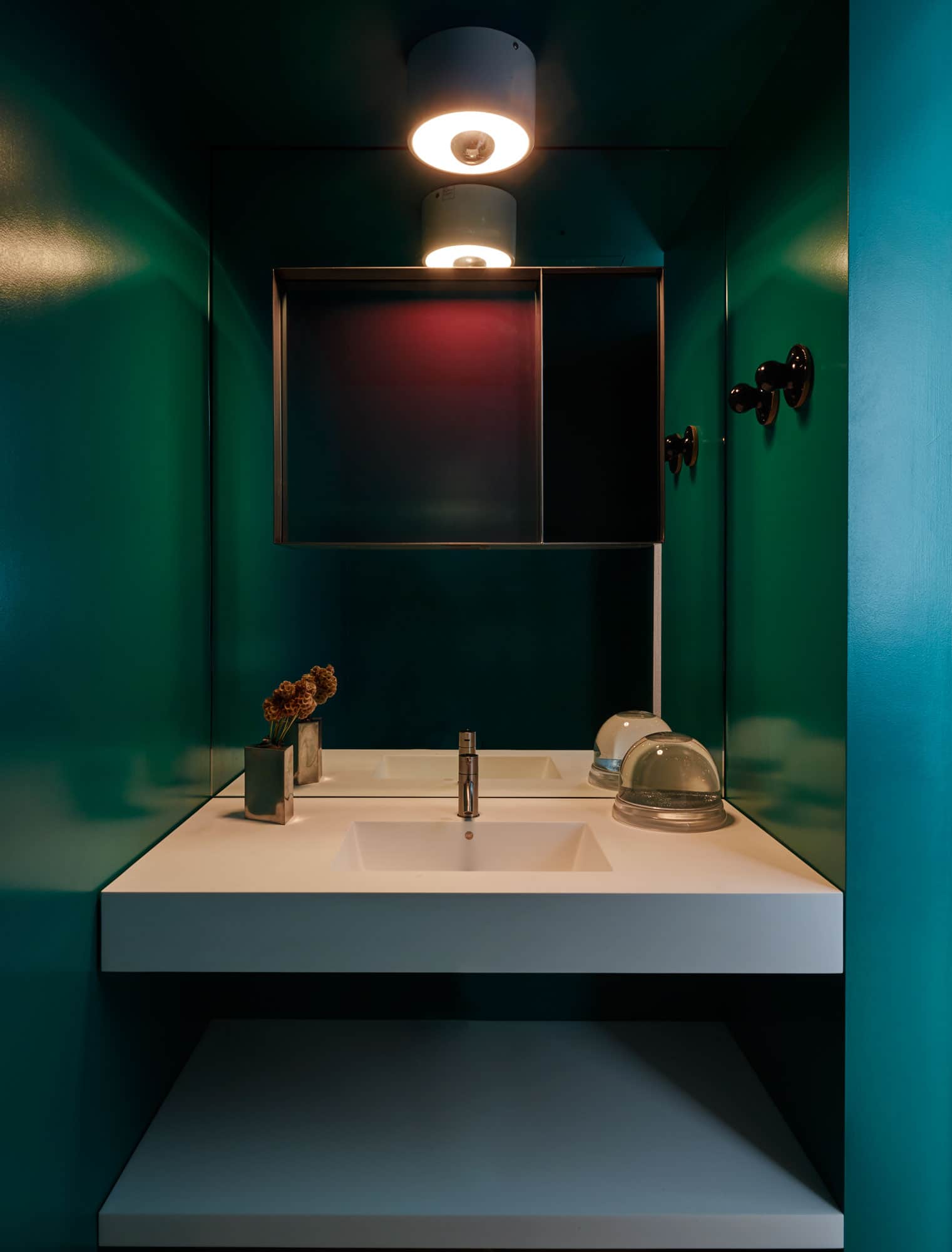 Designed by Carol Egan, this image is a detail shot of a color blocked powder room in a dramatic forest Green painted finish.  There is one source of light shown reflected in the fragmented mirror by Françoise Turner-Larcade.  The light fixture is a flush mount Light by Cini Boeri.  There is a white Corian sink vanity which contrasts the green walls and ceiling. The black ceramic hooks for hand towels are made by M. Crow