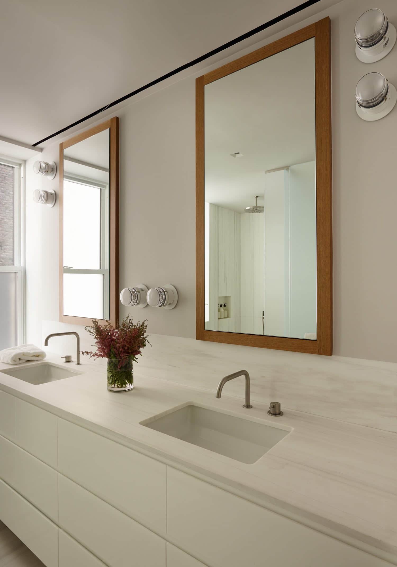 This image shows a double vanity in a Master Bathroom designed by Carol Egan with Fresnel wall lights by Joe Colombo above the sinks and in wall medicine cabinets.  The white marble countertop sits above lacquered white drawer cabinets below.  The faucets are from Vola and are shown in a stainless-Steel finish.