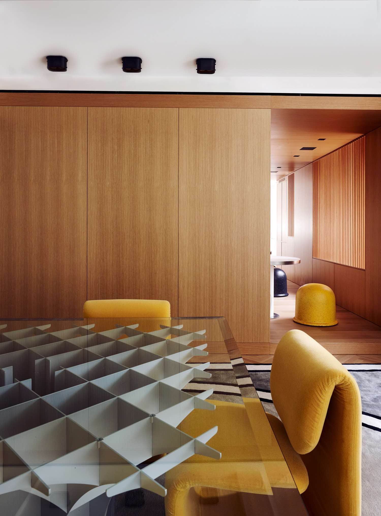 This image shows a dining room designed by Carol Egan featuring the "Cathedrale" table by Pierre Paulin, a sculptural aluminum and glass dining table with the "Programme 1500" chairs by Etienne-Henri Martin from upholstered in yellow wood mohair fabric.  In the background is a paneled oak wall with Flush mount, bronze ceiling lights on the ceiling above.