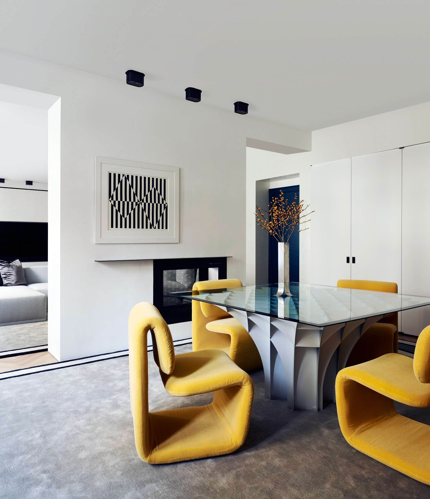 Shown in this image is a view of the dining room designed by Carol Egan featuring the "Cathedrale" table by Pierre Paulin and "Programme 1500" chairs by Etienne-Henri Martin upholstered in a yellow wool mohair fabric by Claremont fabrics.  The artwork above the fireplace is a Silkscreen: "Black & White" by Carmen Herrera.  The area rug is a design by Eric Schmitt Studio.