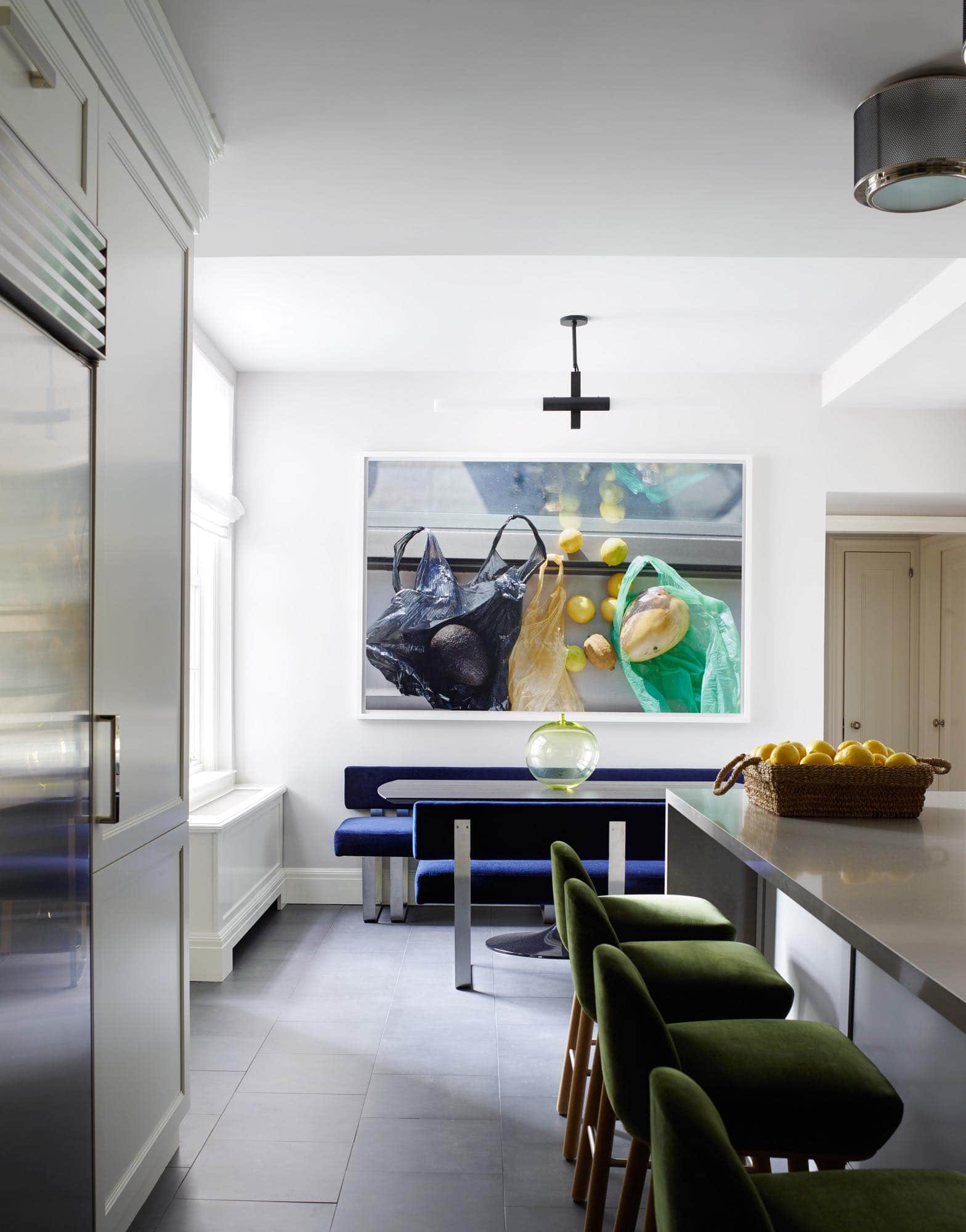 Designed by Carol Egan, shown here is a kitchen in a residence in a Sutton place apartment with Jamie Hayon Beetley style barstools in Racing green Spencer velvet by Claremont fabrics.  The custom brushed nickel mesh ceiling fixture above the kitchen counter is designed by Carol Egan.  On the far wall are custom banquettes in Narcisco cobalt Fabric by Holland & Sherry flanking an ebonized breakfast table with an Applet, Apple vase by Ingeborg Lundin.  The area is lit by a tubular black and white light fixture. above the banquette on the back wall is a photograph by Wolfgang Tillmans.