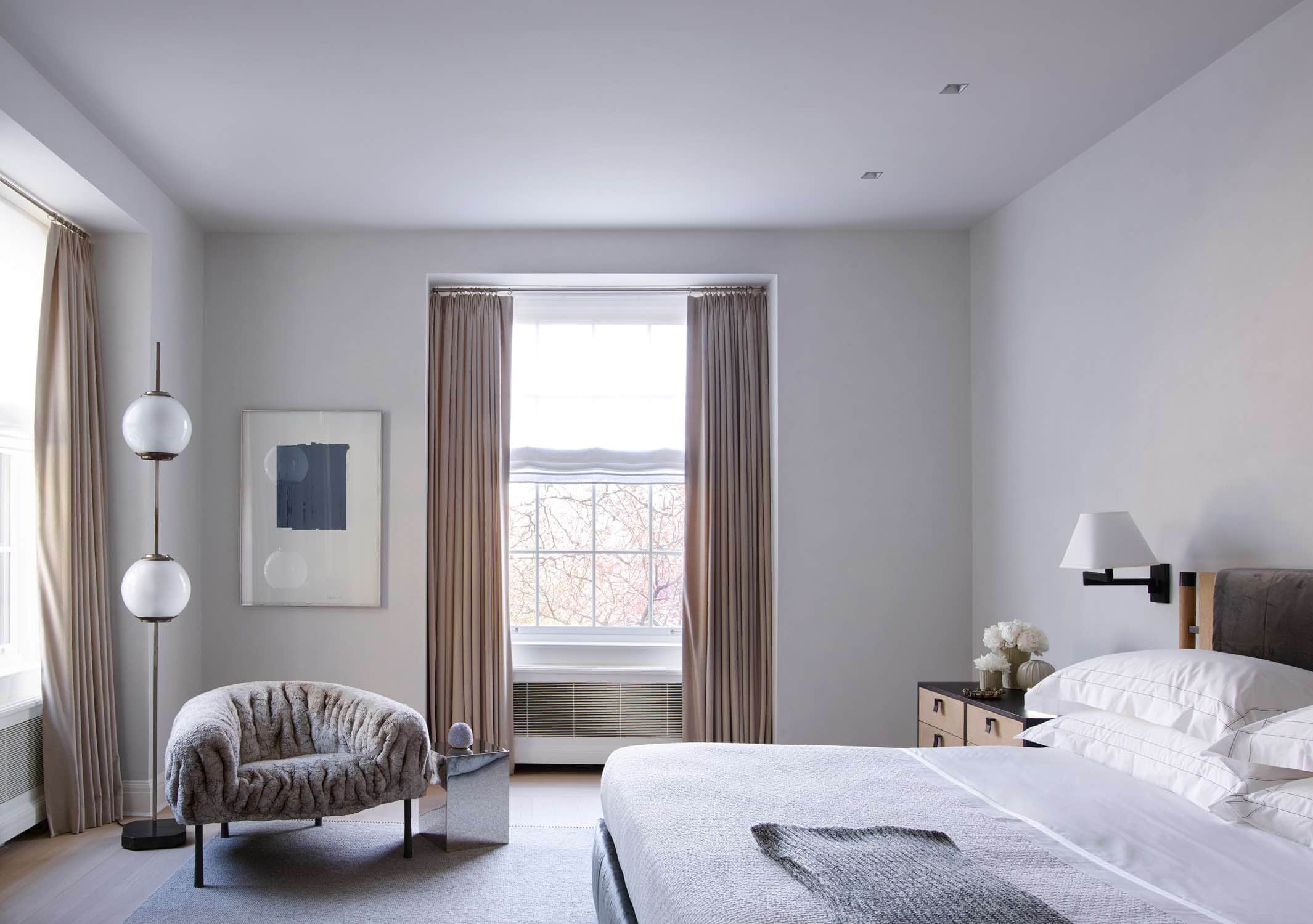 Shown in this image is a master bedroom designed by Carol Egan in this Sutton place residence which showcases a Kuramura Armchair by Ayala Serfat on a custom Sardinian carpet by FJ Hakimian.  The walls are plastered in a pale grey venetian plaster and camel wool tailored curtains hang on Blackened Bronze hardware.