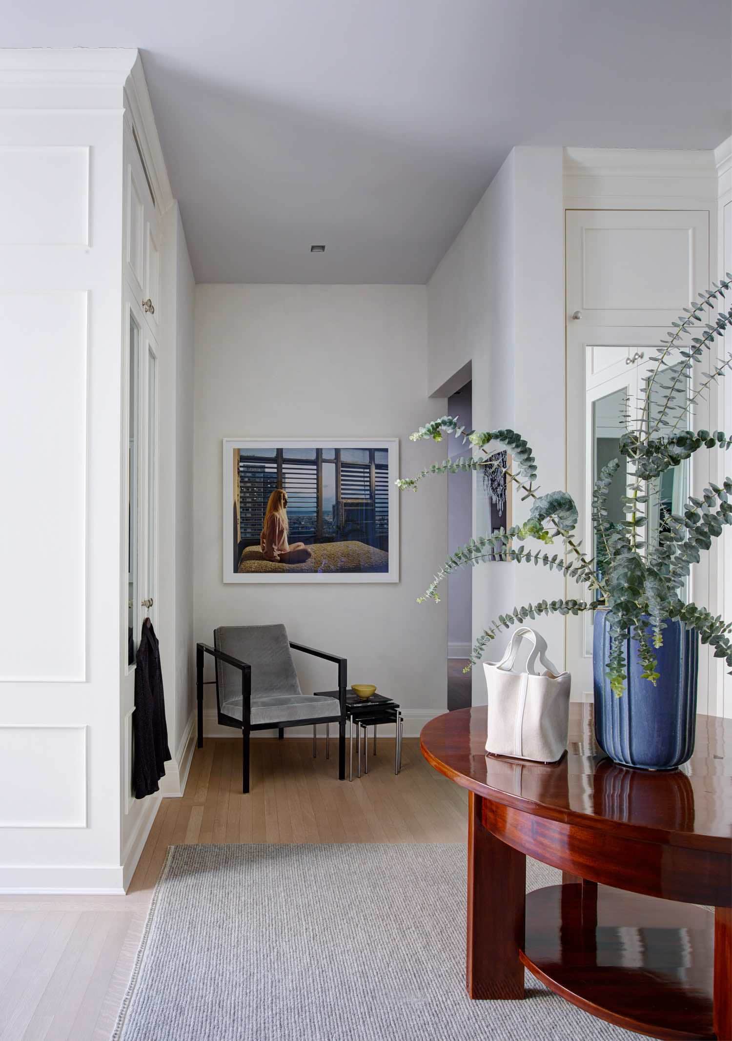 Shown in this image is a view of the master dressing room in this Sutton Place residence designed by Carol Egan.  A Vintage 1930s gueridon Swedish rosewood table showcasing a ceramic vase by Arne Bang floats in the center of the painted millwork which lines the dressing room.  In the corner of the room, an armchair by Michael Pohu with a black metal frame and upholstered seat sits below a framed photograph.  Custom Sardinian carpet by FJ Hakimian.