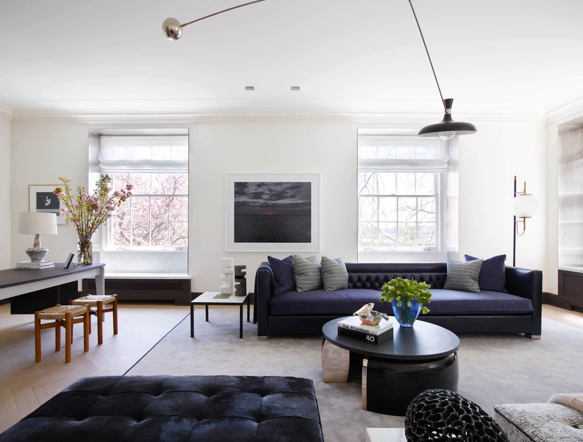 This is an image of a living room designed by Carol Egan.  A Dust Series artwork by Nadav Kander hangs above a custom sofa design by Carol Egan in Navy blue leather and silk velvet upholstery.  The Simple daybed by Charlotte Perriand and double day bench in the style of Pierre Paulin sit opposite the sofa, in front of a Bracelet coffee table by Eric Schmitt.  A mobile light by Fedele Papagni hangs on ceiling.  The window treatments are in Broughton white fabric by Holland & Sherry and the area Rug by V’Soske.