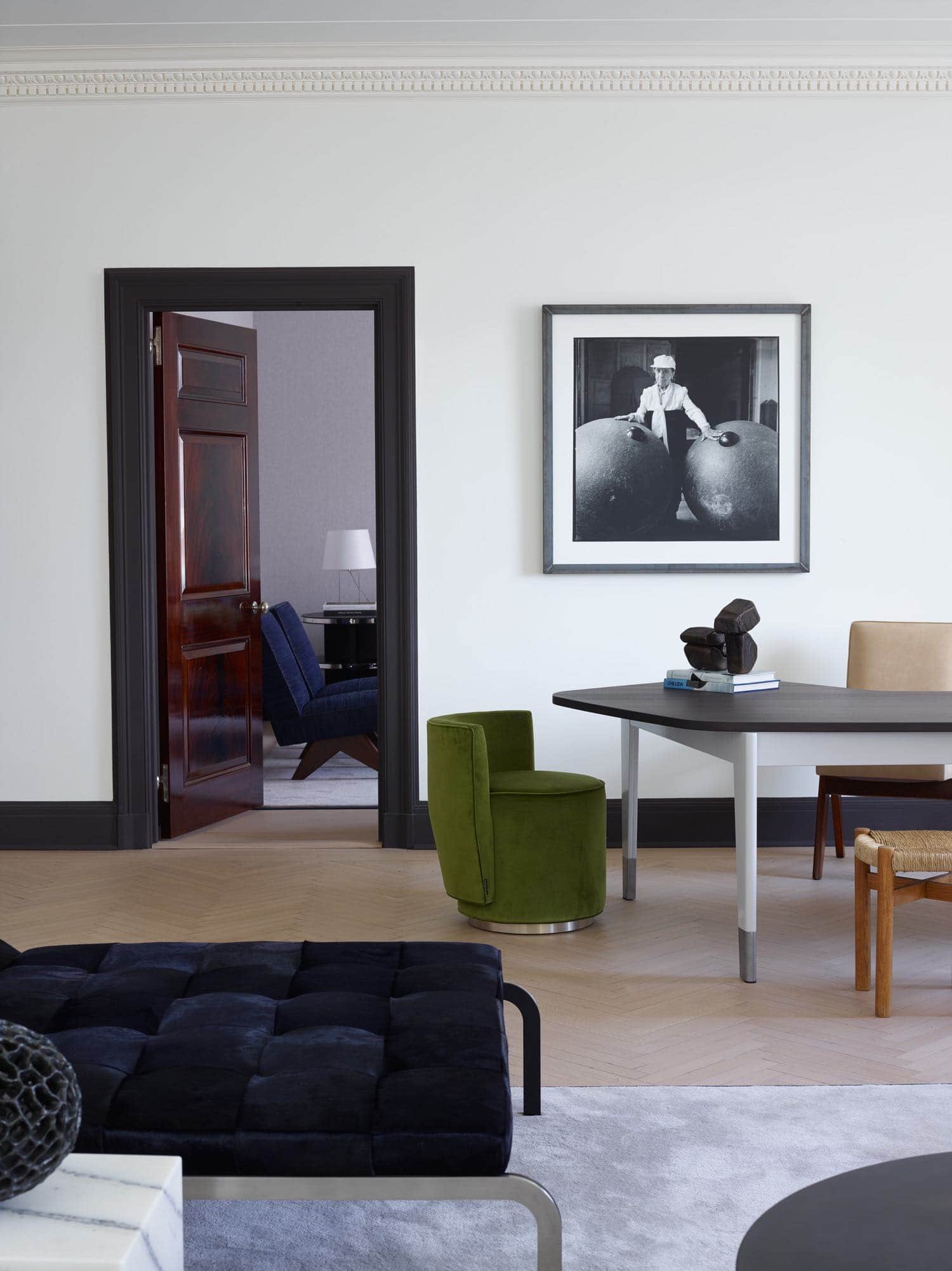 This photograph shows a door leading to the library, French polished and crotch Mahogany doors are contrasted by a charcoal painted door jamb.  Adjacent is a desk by Jean Prouvé flanked by an Oak and Rush Méribel stools by Charlotte Perriand, Bond Street chair by Yabu Pushelberg and Leather armchair by Pierre Jeanneret.  Accessories include ceramic lamp by Mado Jolain and sculptures by Zigor.  Artwork 1995 "Nos Amis" by Jean Francois Jassaud.