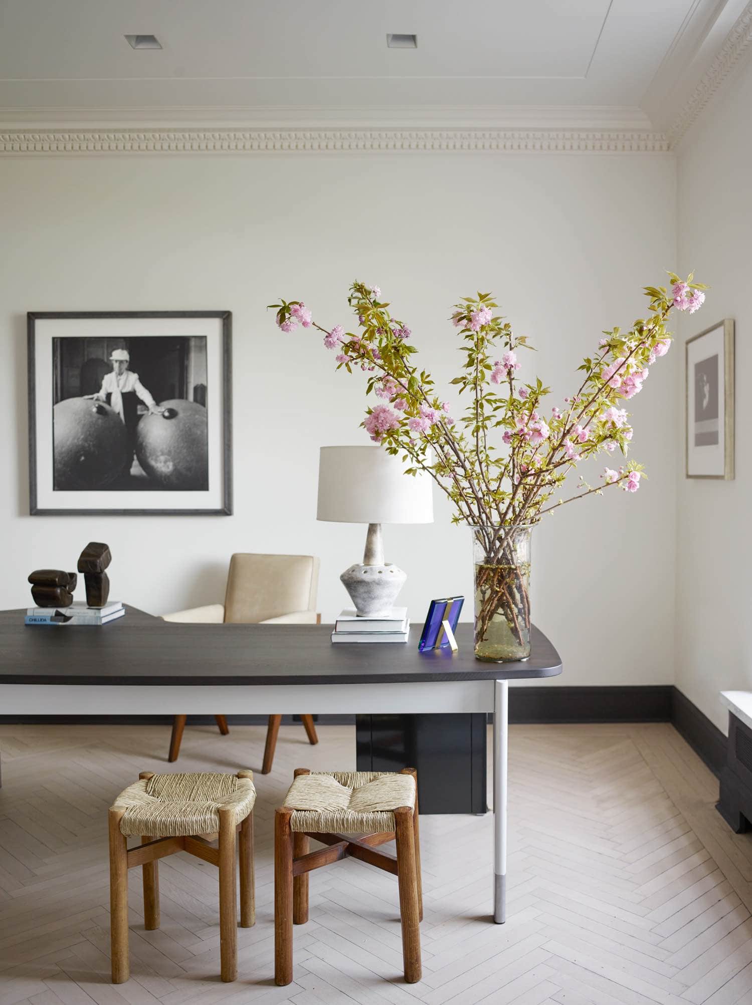 This image shows a desk by Jean Prouvé displaying a ceramic lamp by Mado Jolain and sculptures by Zigor with a Bond Street chair by Yabu Pushelberg, Oak and Rush Méribel stools by Charlotte Perriand and a Leather armchair by Pierre Jeanneret surround it.  On the wall hangs 1995 "Nos Amis" artwork by Jean Francois Jassaud in this Sutton Place residence designed by Carol Egan.