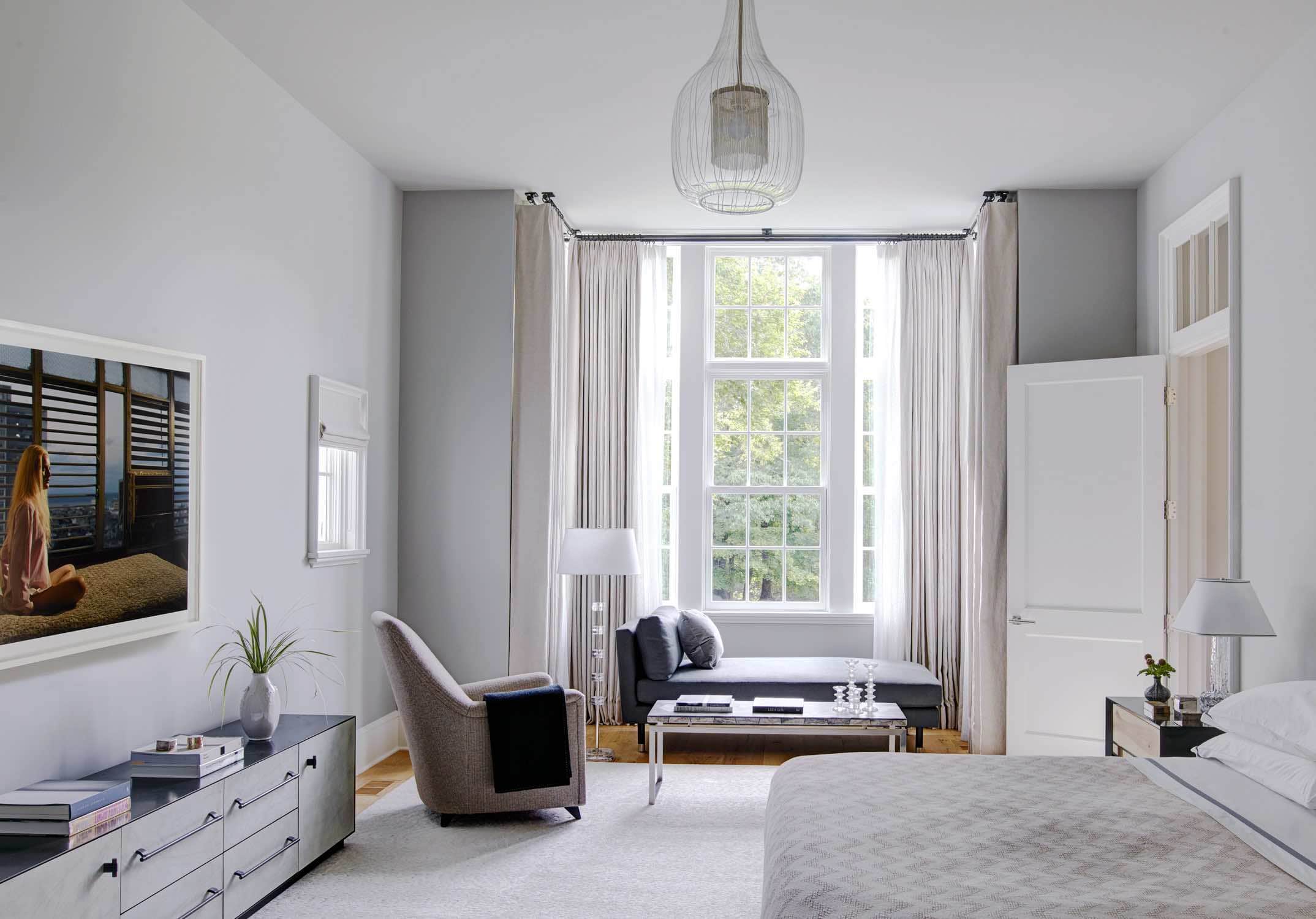 An image showing a master bedroom designed by Carol Egan.  The headboard is by Roman Thomas. The bed is dressed in linen bedspread by Matouk linens.  Opposite the bed is a console by BDDW, and a French Deco armchair from the 1930s is from Galerie Blanchett in Paris.  By the window is a 1960 floor lamp by Carl Fagerlund for Orrefors, a custom chaise with exposed wood & nickel sabots by Doreen Interiors in Burnished Canvas and a Vintage marble top & nickel base coffee table.  The room is lit by a 1970s ceiling light by Angelo Brotto for Esperia.  The rug is by Mitchell Denberg and the walls are painted in pavillion gray by Farrow & Ball.