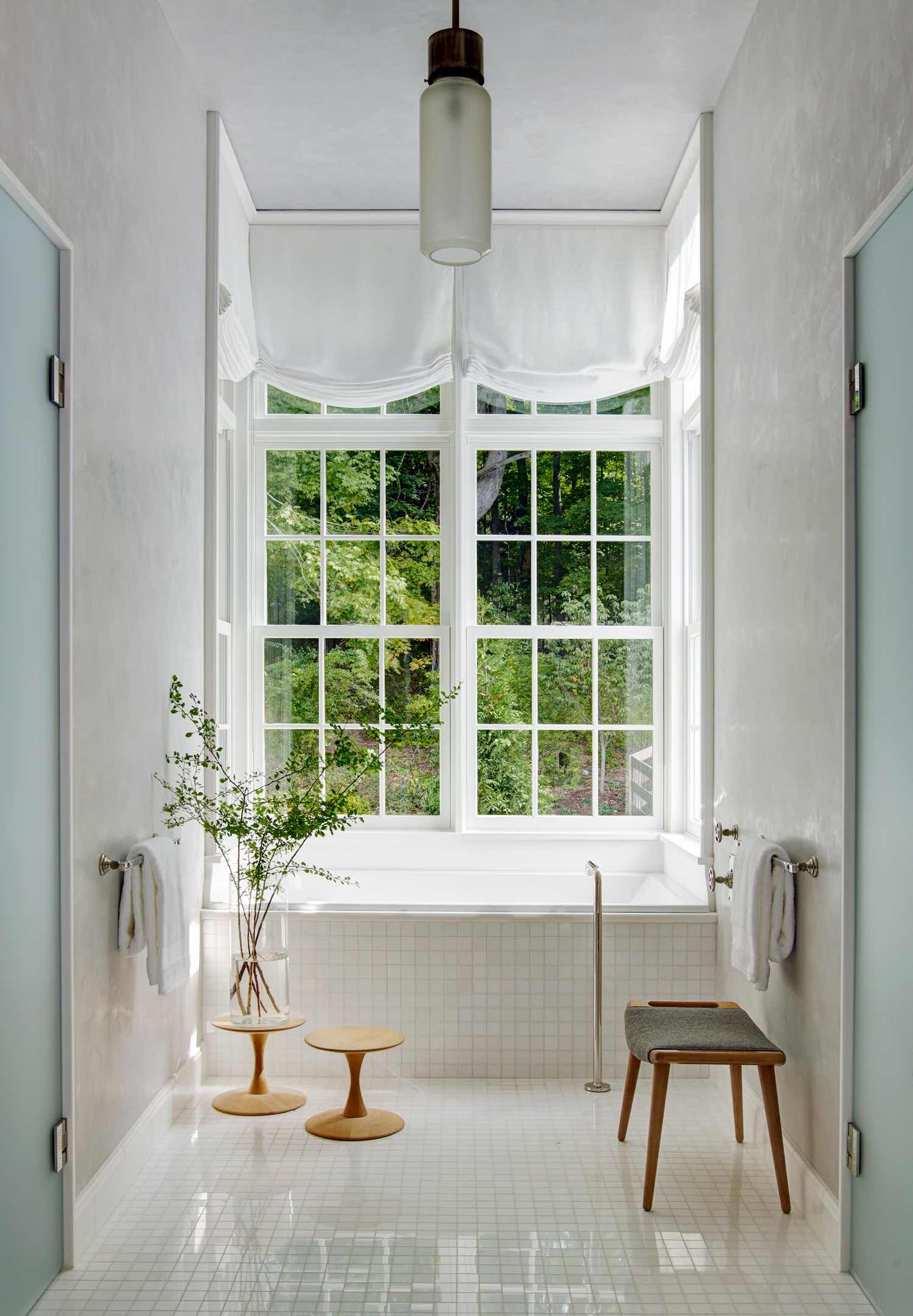 Designed by Carol Egan, the master bathroom floors, and bathtub apron are tiled white Thassos mosaic with fittings by Waterworks.  Adjacent to the bathtub is a Hans Wegner stool upholstered in grey wool and small childs stool by Nanna Ditzel holds a vase of Fresh cut branches from the garden.  1970s Luigi Caccia pendant light hangs from the ceiling and the walls are sheathed in Venetian plaster.  The bathtub is framed by a large window overlooking the garden meadow.