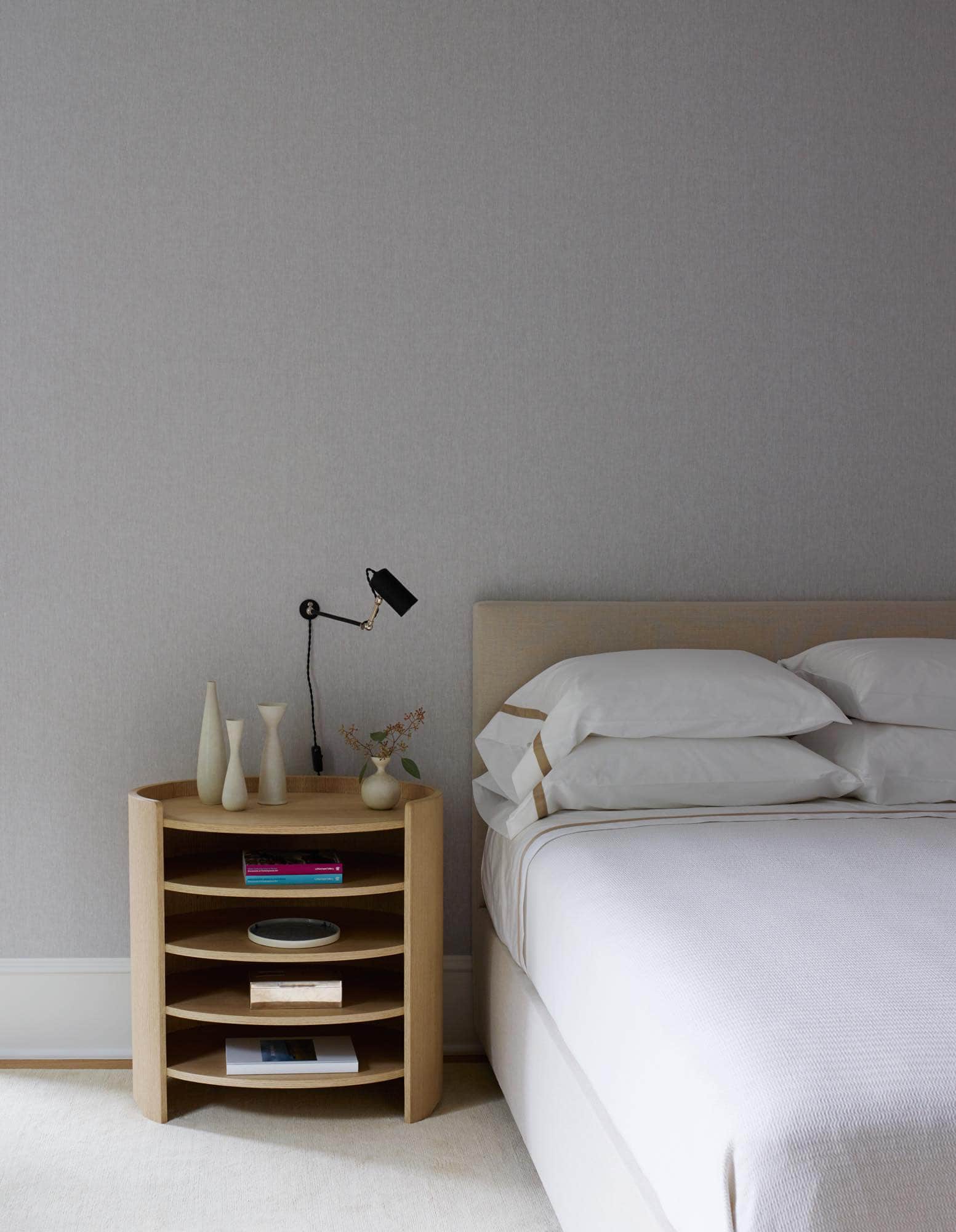 This image shows a guest bedroom consisting of a custom bed and oval night tables with five shelves designed by Studio Carol Egan.  Plugin wall lights from Alain Fradin are installed on walls in Holland & Sherry Alona Rice paperbacked linen fabric by Holland & Sherry.  Pillows in Tendril fabric by Claremont and floor covering in hand-knotted wool area rug by Sam Kasten.