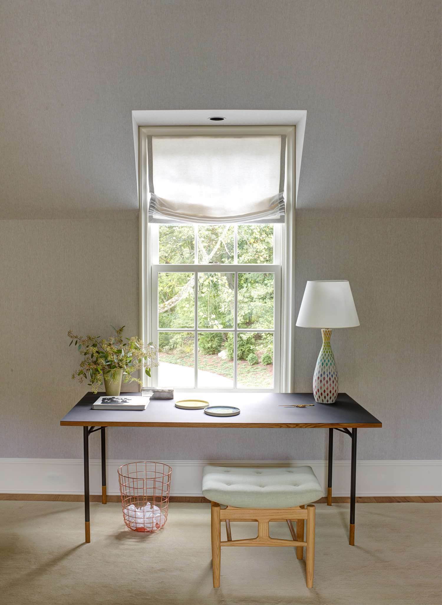 An image showing a study designed by Carol Egan.  A writing table by Finn Juhl by the window with a stool by House of Finn Juhl and a Baton desk lamp by Lightolier.  The walls are upholstered in a natural linen and the desk is set under the window which overlooks the garden views from the 2nd floor.