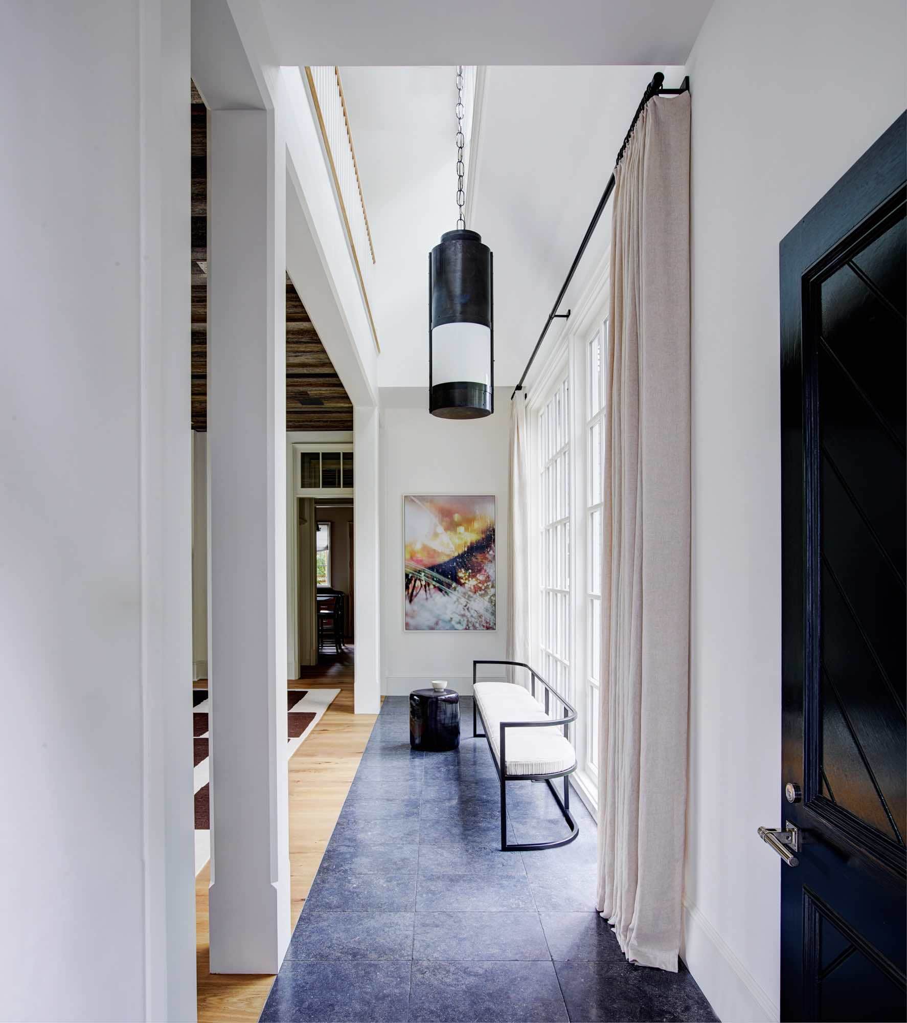 Shown here , the foyer of a Greenwich residence designed by Carol Egan, the Front door is a painted herringboned pattern in gloss ebony finish with custom hardware by Nanz in a polished Nickel finish.  The Foyer floor is large slabs of Belgian Blue Stone where a metal and upholstered bench by Lucca & Co is upholstered in hand-woven Ottoman Stripe/Broken Ikat fabric by Sam Kasten.  A Vintage Itlaian Lantern is hung from the ceiling in blacked metal and opaline glass and there are tailored linen curtains framing the window.  Artwork by Marilyn Minter hangs in the Foyer.